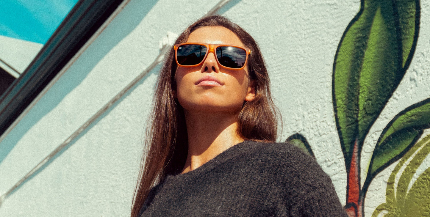 A confident woman with long brown hair stands outside in sunlight, wearing Neven Eyewear's Suggestion orange frame sunglasses. She gazes upward before a white wall with a green leaf mural and part of a building roof behind her.
