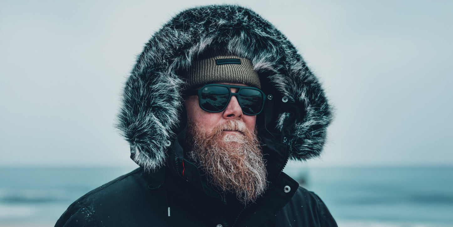 A bearded person outdoors wears a dark parka with a fur-lined hood, Neven Eyewear Cosmo aviator sunglasses featuring polarized UV400 protection, and a knit beanie against a cold, snowy waterside landscape under an overcast sky.