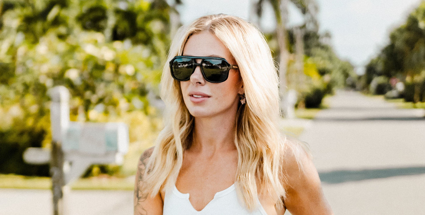 A woman with long blonde hair wears Neven Eyewear's Cosmo aviator sunglasses and a white tank top, standing outdoors on a sunny day. Lush greenery and palm trees line the sunlit street behind her, highlighting the Cosmo collection.