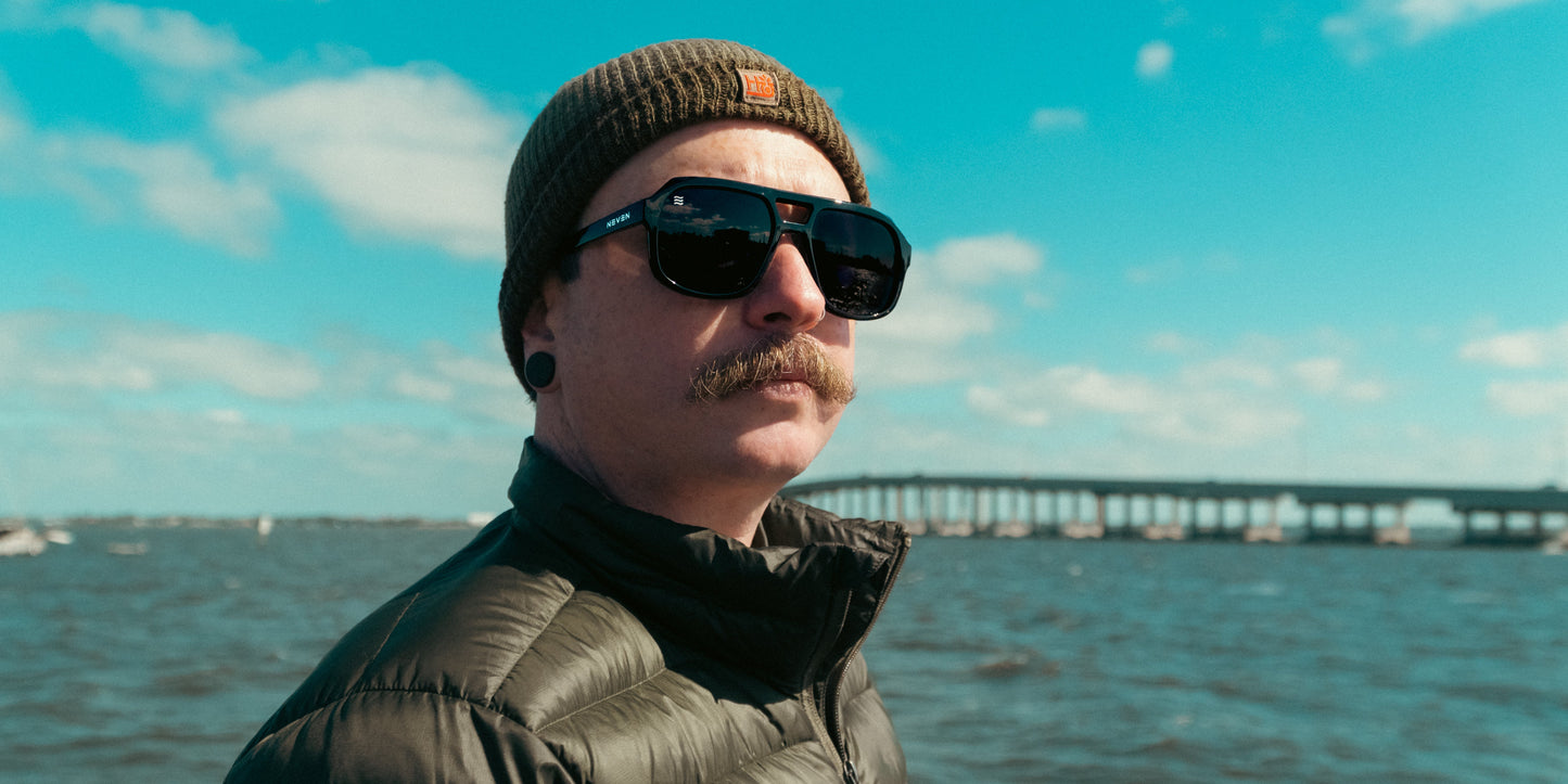 A man with a mustache wears Neven Eyewear’s Cosmo aviator sunglasses featuring polarized UV400 protection, a green knit beanie, black ear gauges, and a green puffer jacket by the water, with a long bridge and blue sky in the background.