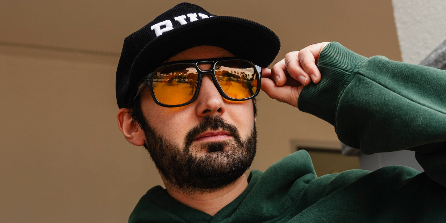 A man with a dark beard wears a black cap and dark green hoodie as he adjusts his Neven Eyewear Helios aviator sunglasses with yellow, UV400 lenses, gazing into the distance against a neutral, blurred background.