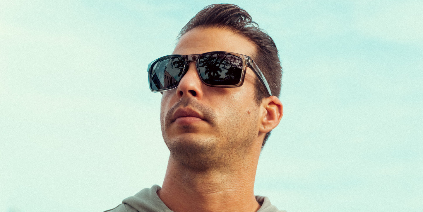 A man with short dark hair wears Neven Eyewear's Breakers XL sunglasses for large heads and a light gray hoodie. He looks upward, tree reflections visible in his lenses, set against a clear pale blue sky.