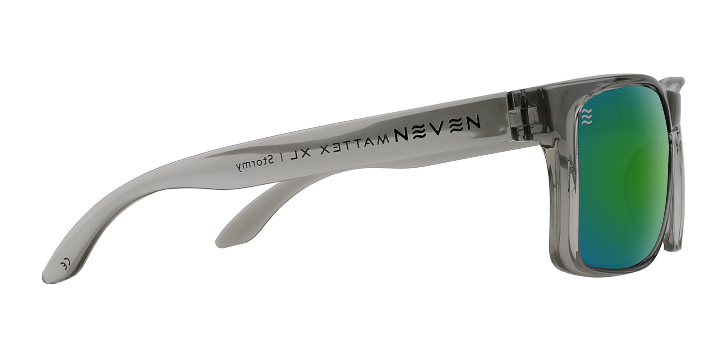 A pair of oversized, chunky Stormy XL sunglasses by Neven Eyewear is shown from the side. The bold rectangular green mirrored lenses reflect light, and the transparent gray frame features arms labeled “NEVEN MATTE XLI Stormy.”.