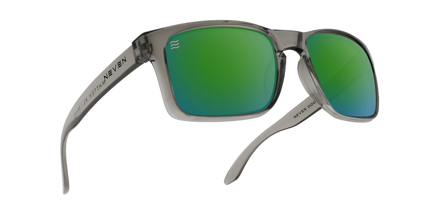 The Stormy XL by Neven Eyewear are oversized translucent gray rectangular prescription sunglasses with green mirrored polarized lenses. The temples display NEVER ORDINARY and a wavy line. Shown angled on a white background.