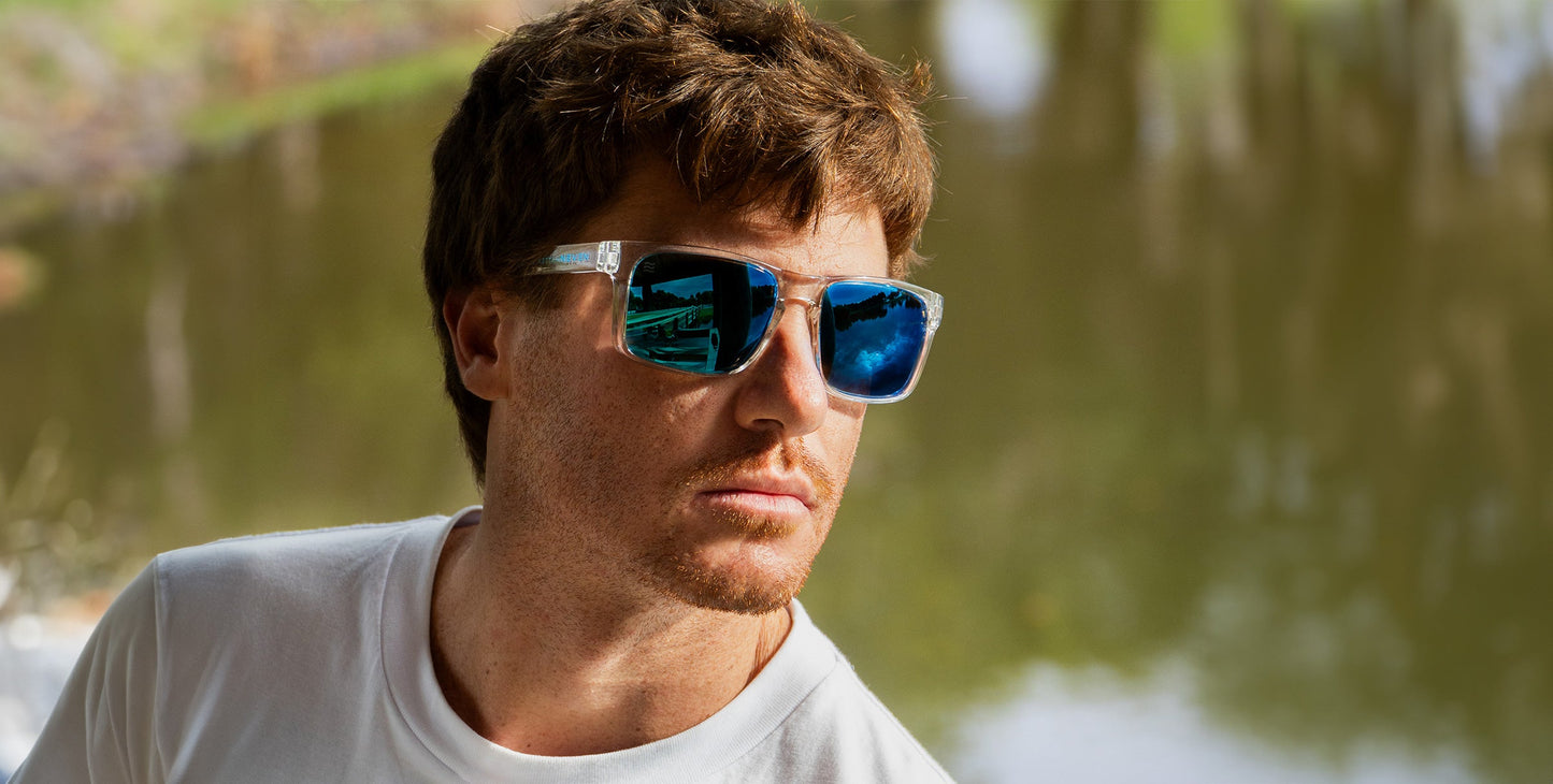 A man with short brown hair and light stubble wears Neven Eyewear Surge XL blue-mirrored, polarized sunglasses and a white T-shirt, standing outdoors near water with trees and grass reflected in his lenses.