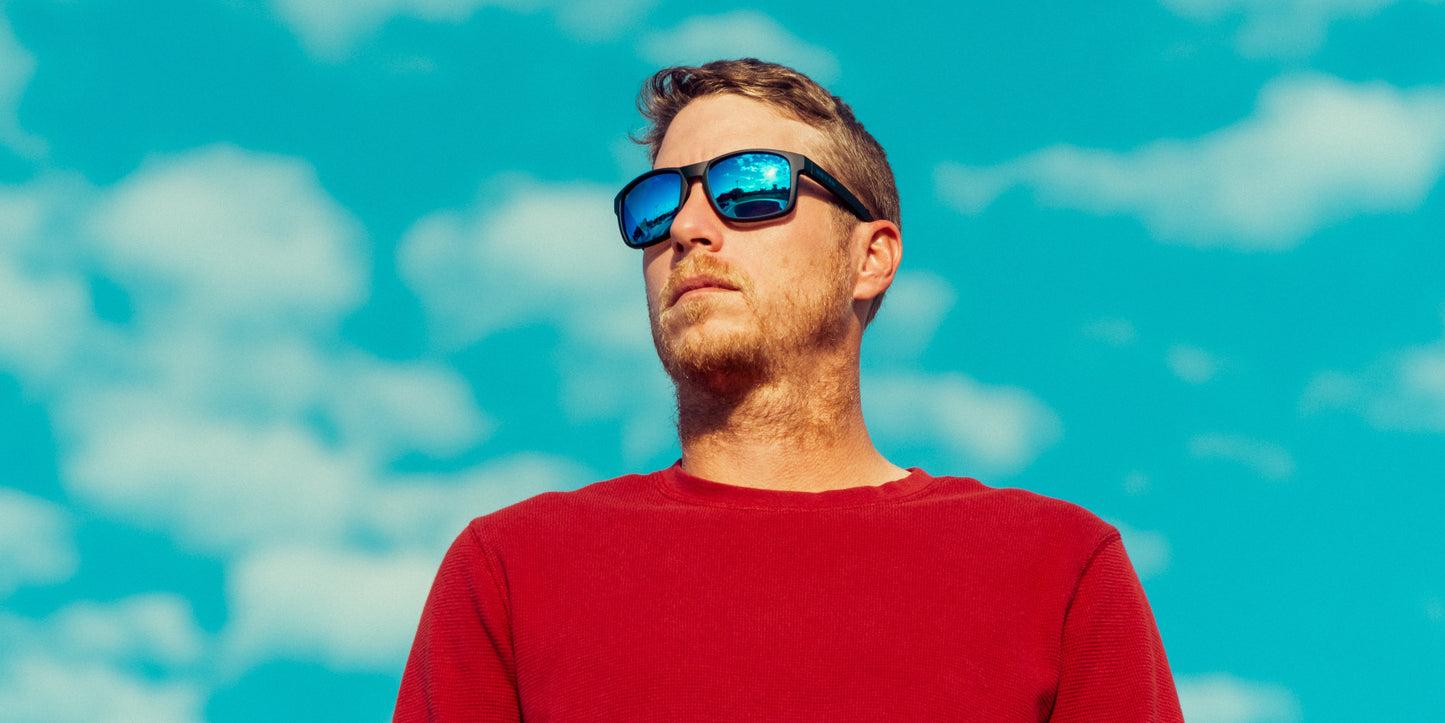 A man in a red long-sleeve shirt wears Neven Eyewear’s Blue Steel polarized sunglasses with UV400 protection, standing outdoors under a bright blue sky with scattered clouds for a vibrant, summery look.