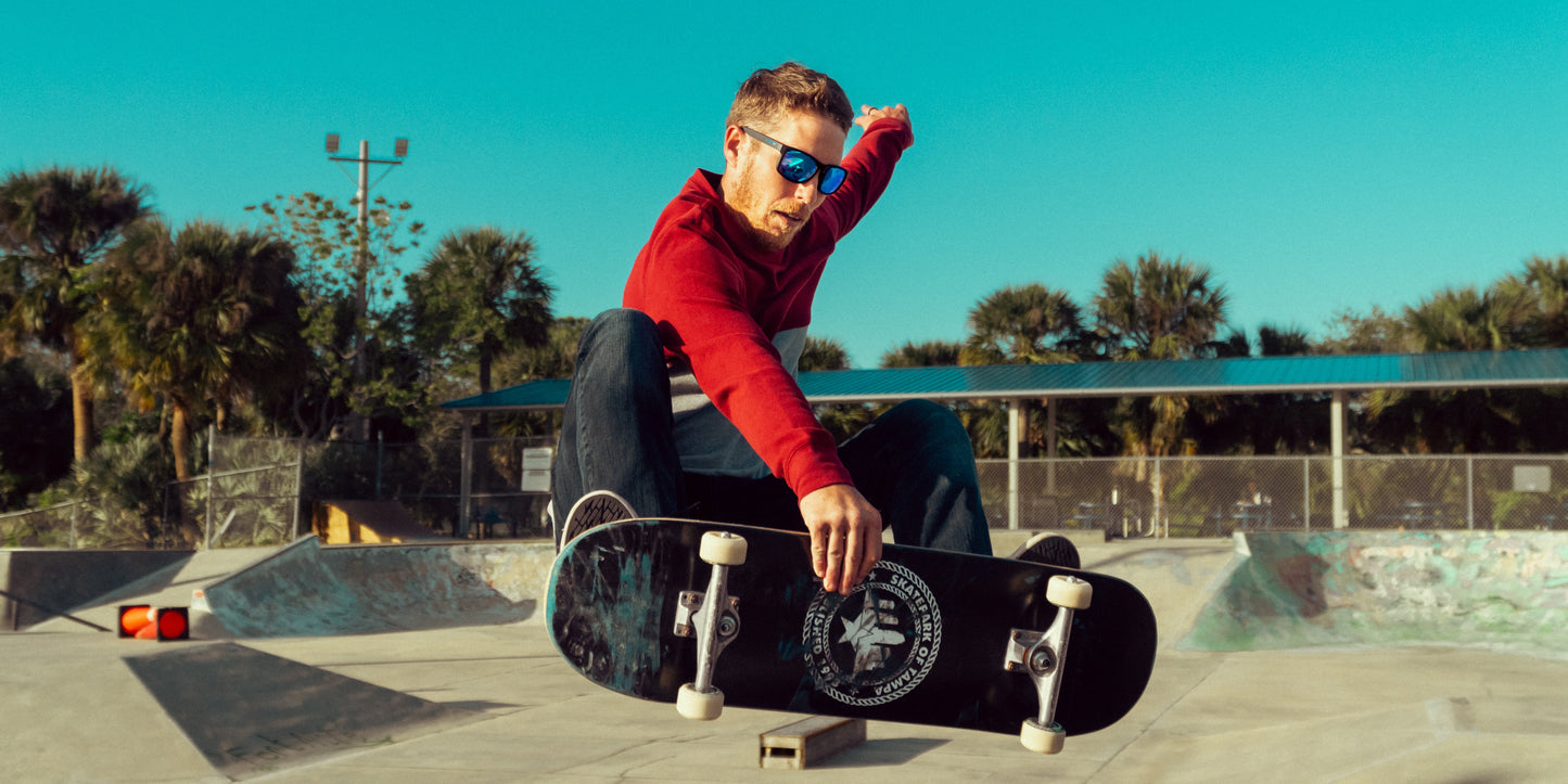 A man wearing Neven Eyewear Blue Steel polarized sunglasses and a red long-sleeve shirt performs a skateboard trick at a sunny skatepark, crouching to grab his board with palm trees and a blue-roofed shelter in the background.