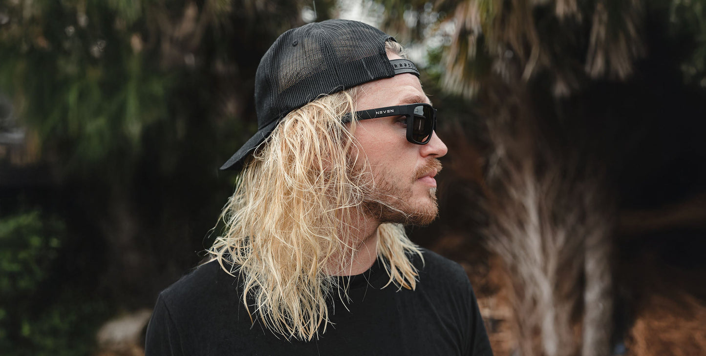 A person with long, wavy blond hair and a beard wears a black t-shirt, a backward black baseball cap, and Neven Eyewear Doom Polarized UV400 sunglasses. The background has blurred greenery and palm trees.