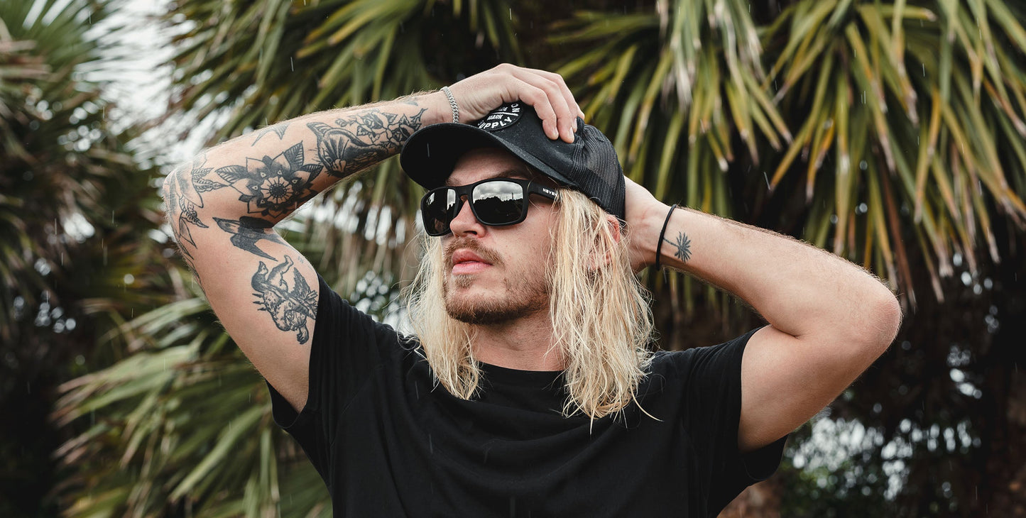 A person with long blond hair, tattooed arms, and a black T-shirt and cap wears Neven Eyewear’s Doom Polarized UV400 sunglasses with impact-resistant lens, standing outdoors by palm trees and touching their cap while gazing into the distance.