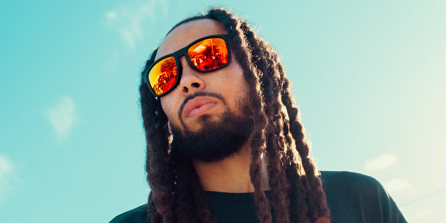 A man with long dreadlocks and a trimmed beard wears Neven Eyewear’s John Wayne sunglasses with mirrored lenses, reflecting a crowd. He looks serious and confident under a bright blue sky, sunlight highlighting his hair and face.