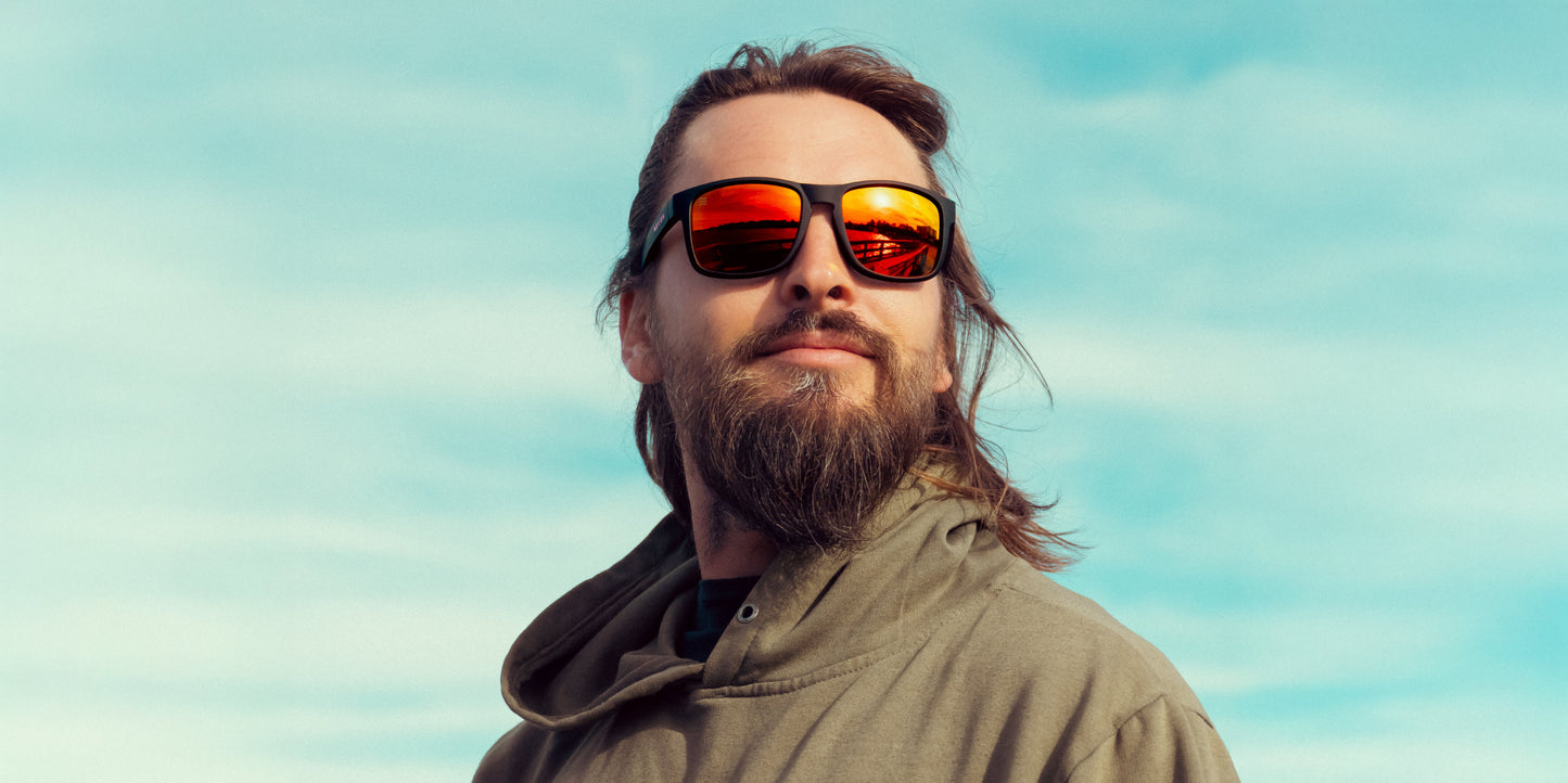 A man with long hair, a beard, and mustache wears a green hooded jacket and Neven Eyewear John Wayne sunglasses with polarized UV400 lenses, gazing sideways. The bright blue sky and sunset are mirrored in his shades.