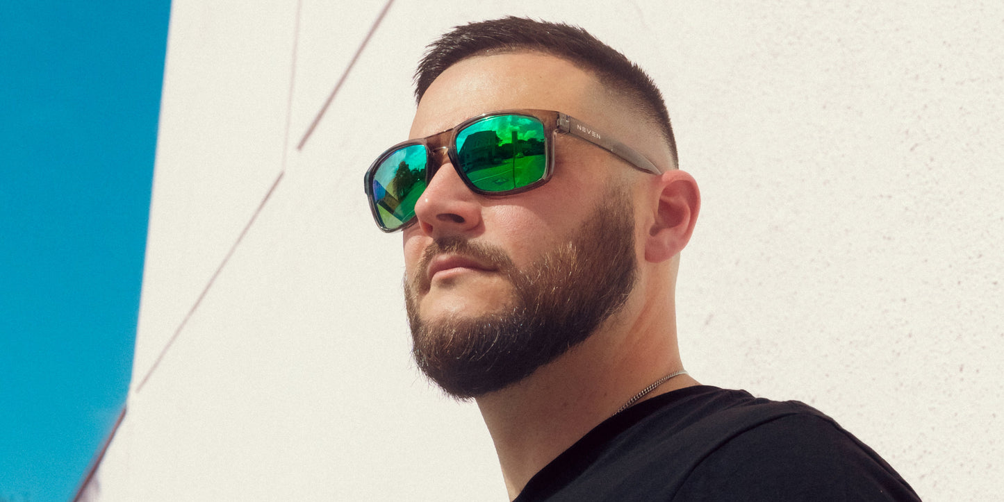 A man with a trimmed beard and short hair wears Neven Eyewear’s Stormy reflective green sunglasses with polarized UV400 protection and a black shirt, standing outdoors in bright sunlight against a white wall and clear blue sky.