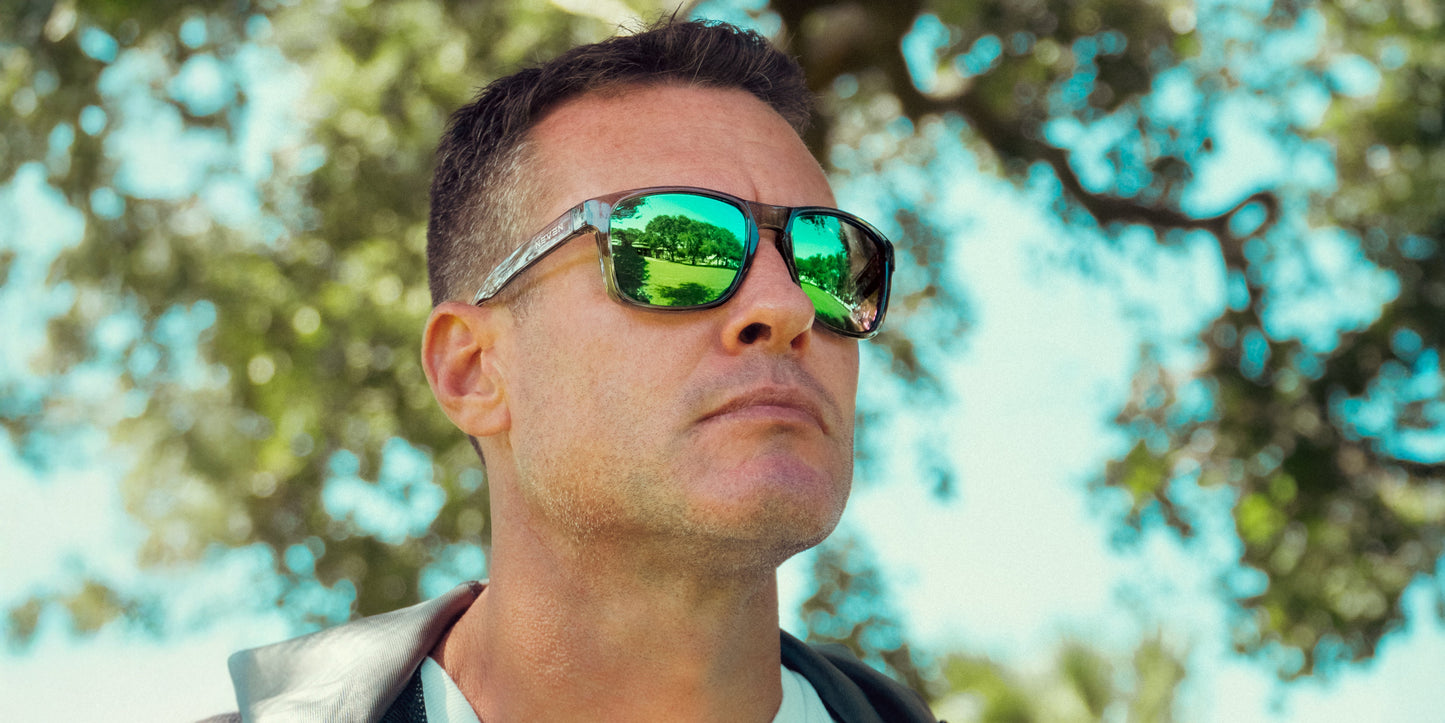 A man in Neven Eyewear Stormy sunglasses with impact-resistant lenses looks up outdoors. Reflections of green trees and blue sky appear on his shades. He wears a light jacket, enjoying the bright, sunny day.