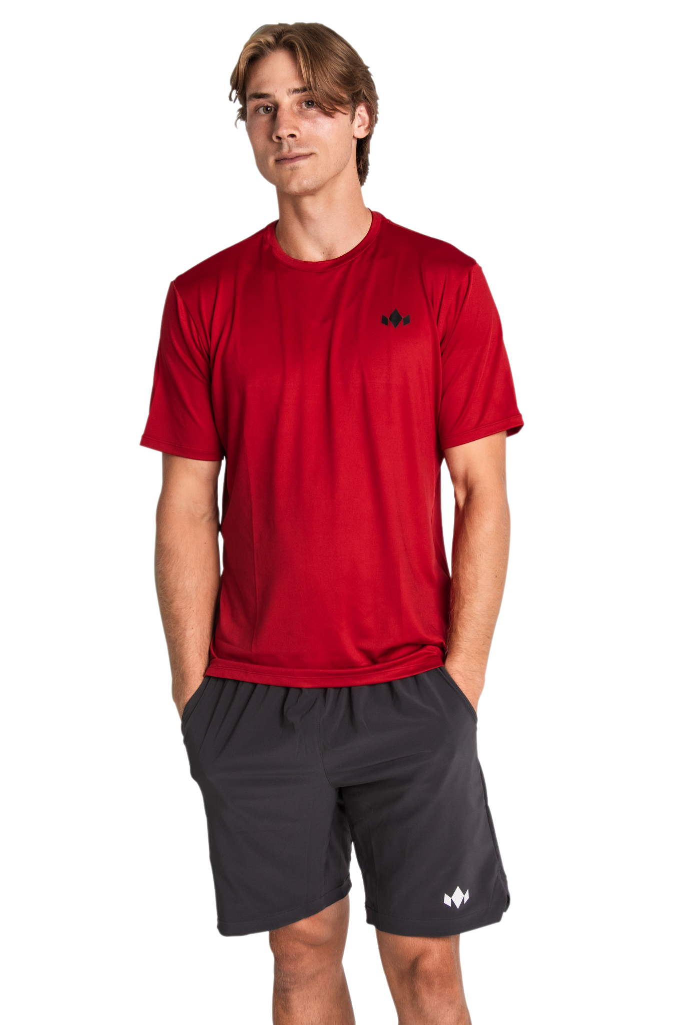 A young man with light brown hair stands against a white background, wearing a Diadem Sports - Men's Essential Everyday Tops red short-sleeve shirt with a black logo and black shorts with a white logo, hands in his pockets.