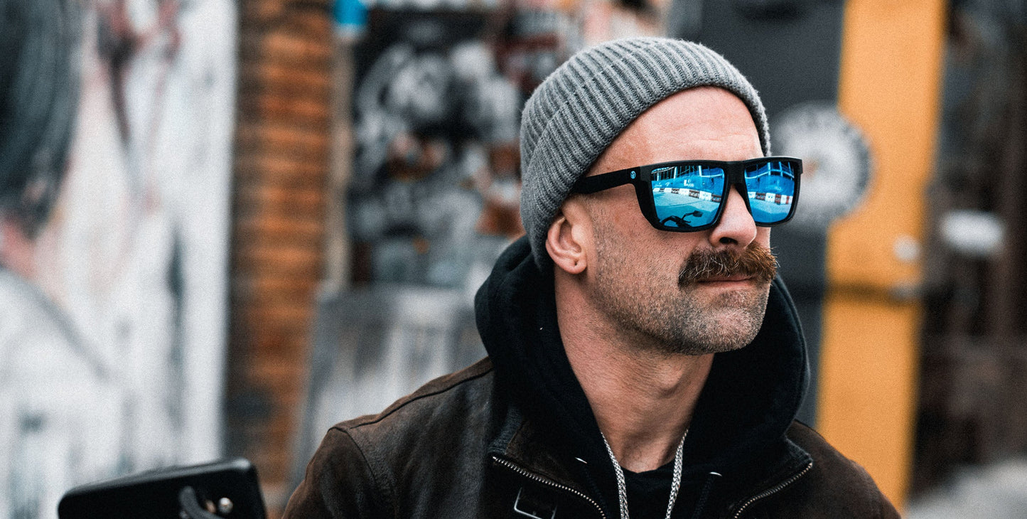 A man with a mustache wears Neven Eyewear's Honest John's polarized sunglasses and a gray beanie, reflecting city streets. He stands outside in front of graffiti walls, dressed in a dark jacket layered over a hoodie.