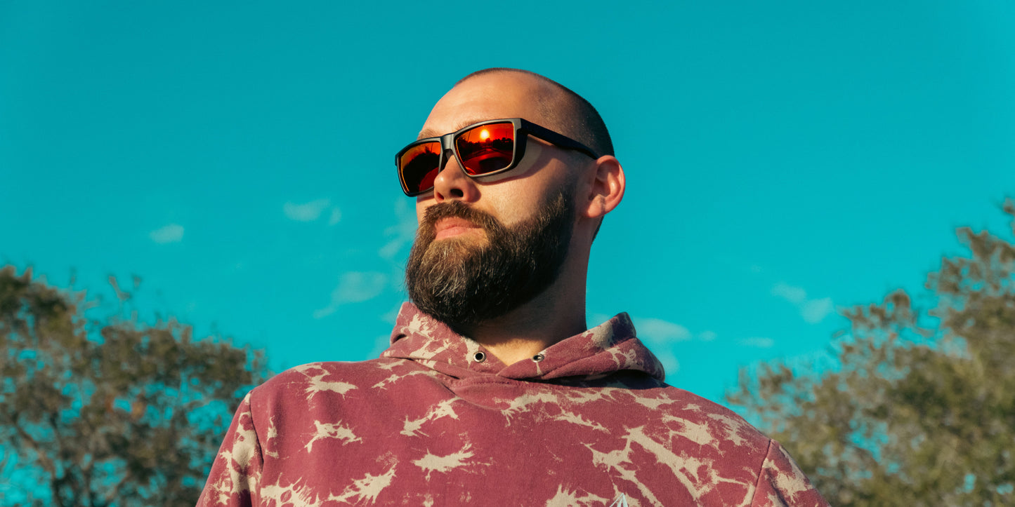 A bearded man wears Neven Eyewear's Long Point sunglasses with polarized UV400 protection and a maroon tie-dye hoodie. Outdoors among trees and a clear blue sky, sunlight reflects off his sunglasses as he gazes into the distance.