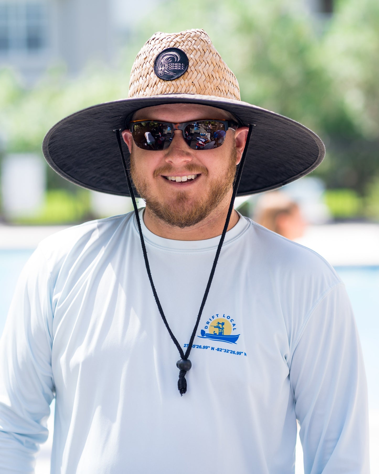 A man sports Victor Sunglasses by Epoch Eyewear, a wide-brimmed straw hat with a chin strap and front logo, and a light blue long-sleeved shirt as he smiles outdoors against a sunlit, green, blurred background.