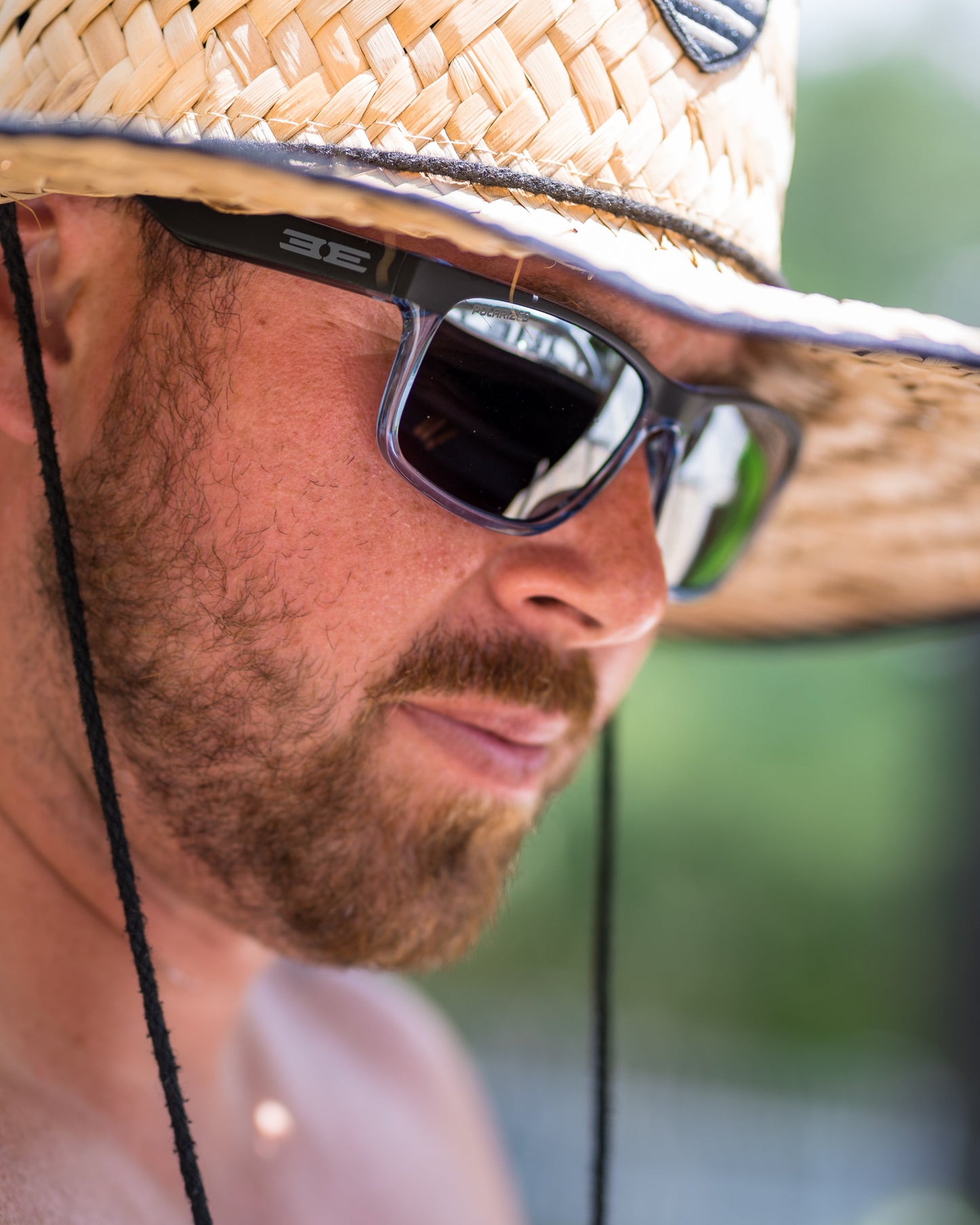 Wearing Epoch Eyewear’s Delta black sunglasses and a wide-brimmed straw hat with black strings, a brown-bearded man stands outdoors on a sunny day, focused against a blurred green backdrop.