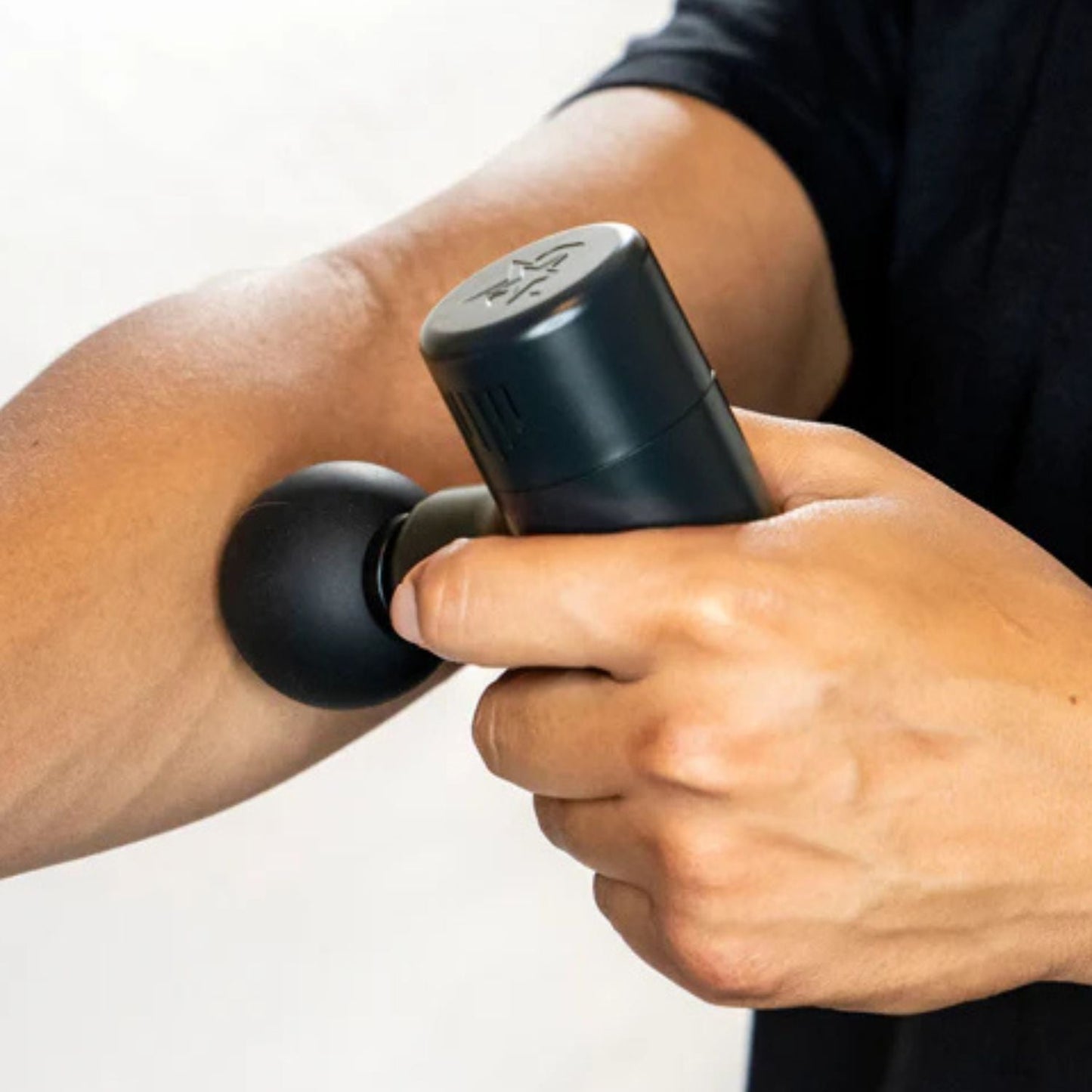 Wearing a dark shirt, a person uses the iReliev MVPmini Percussion Massage System with a round attachment on their forearm to ease sore muscles. The portable massage gun and arm are highlighted against a plain white background.
