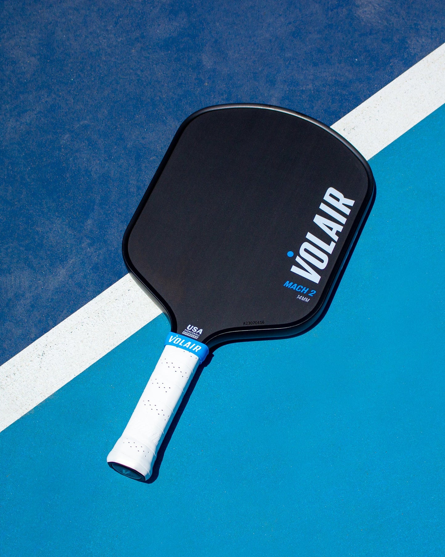 A Volair Pickleball Mach 2 Paddle with bold white VOLAIR branding rests on a blue court, crossing a white boundary line. The handle features white grip tape with blue accents, plus USA and VOLAIR near the end. Money Back Guarantee.