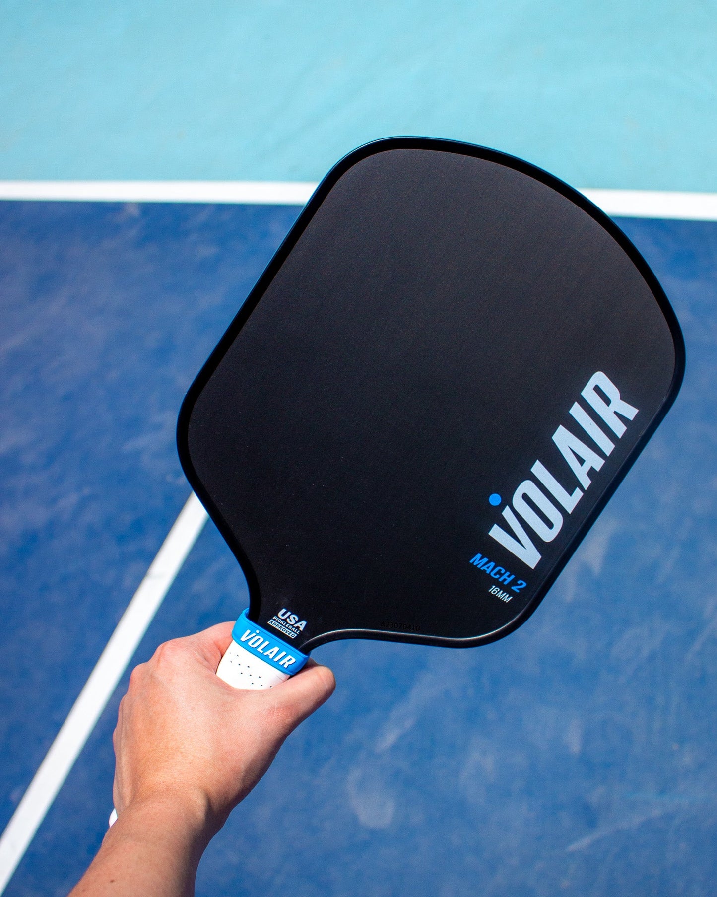 A hand holds a black Volair Pickleball Mach 2 Pickleball Paddle with a white grip over a blue court. The paddle shows “Mach 2” and “USA Pickleball Approved” near the handle. Includes a Money Back Guarantee.