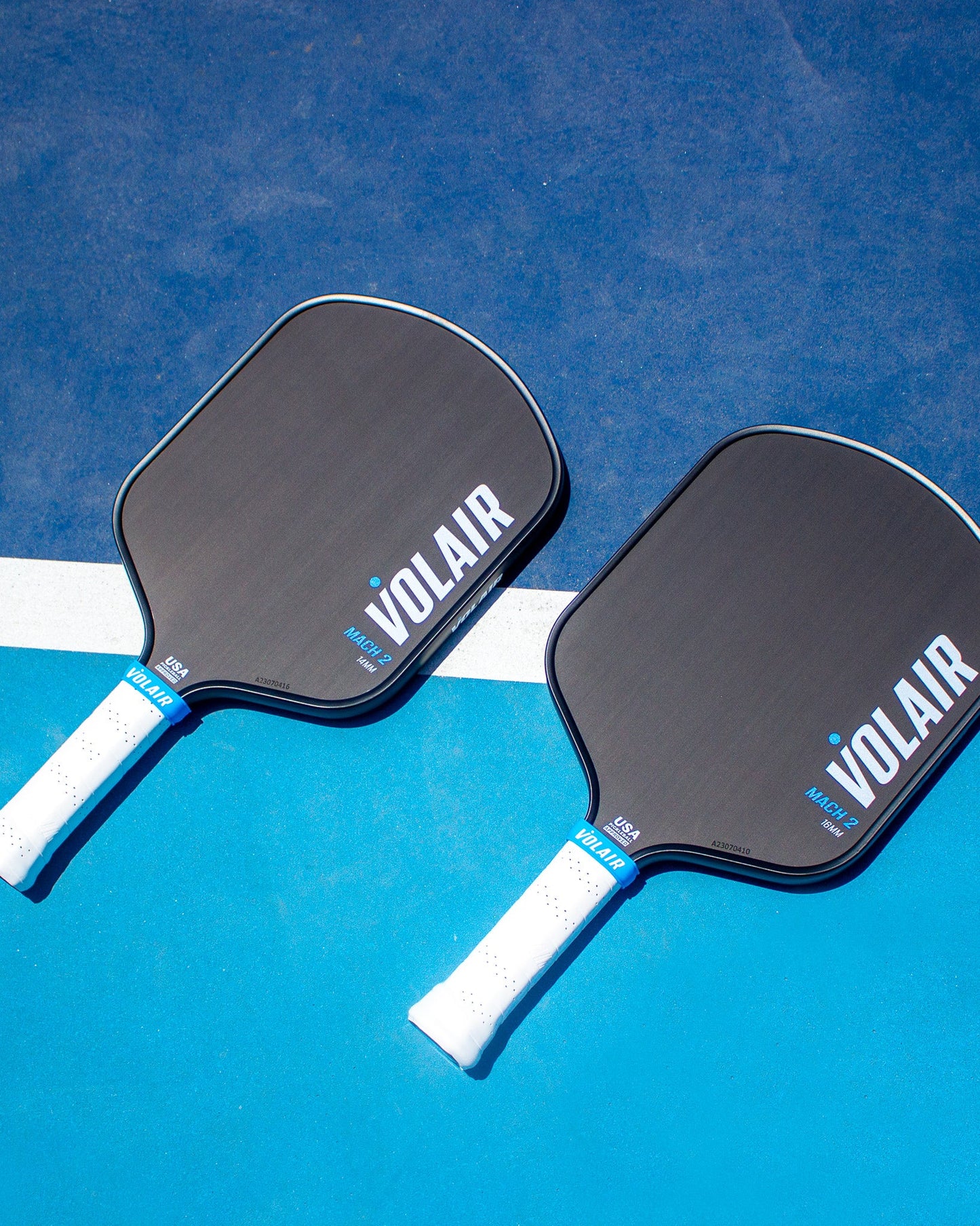Two black Volair Pickleball Mach 2 paddles with white handles rest side by side on a blue outdoor court, crossing a white line. Both display the “VOLAIR” logo. Paddle comparison chart available.