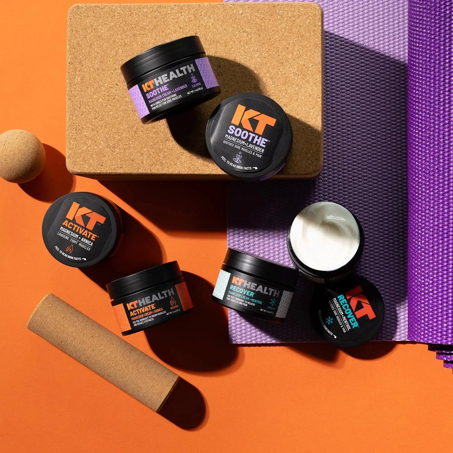Four small KT Tape KT Health Magnesium Creams labeled Soothe, Activate, and Recover sit on a cork yoga block and purple mat. One jar is open, revealing white cream. Cork massage rollers are nearby on an orange surface.
