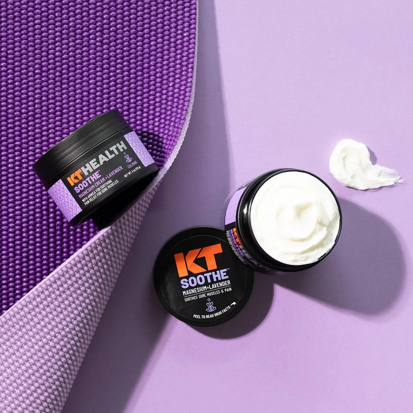 Three black containers of KT Tape KT Health Magnesium Creams are on a purple exercise mat and lilac surface. One is open, revealing white cream inside, with some spread beside it. The containers have orange and white text labels.