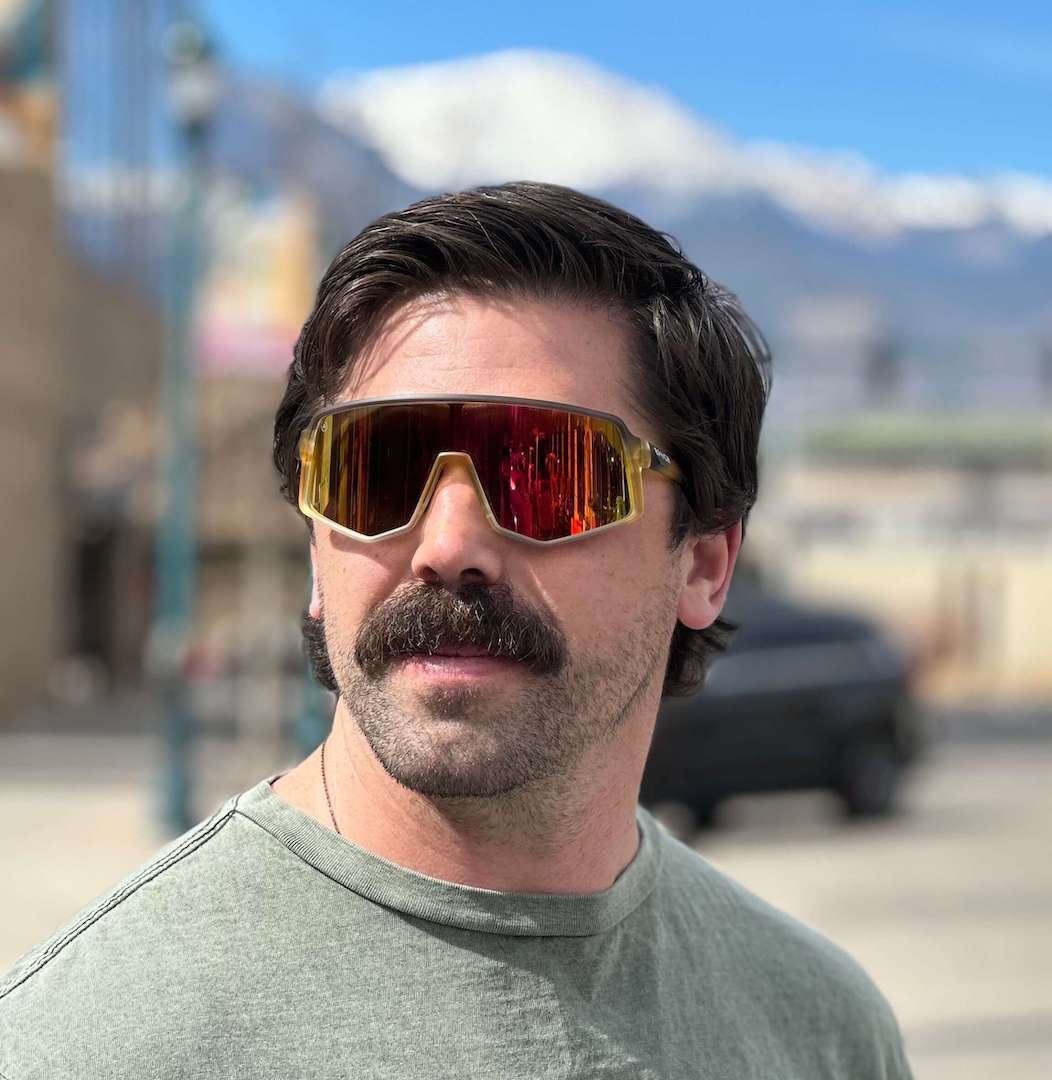 A man with dark hair and a thick mustache wears Epoch Eyewear’s L2 Sport Wrap Sunglasses—Legendary Design for All—featuring a lightweight fit and large red-and-yellow reflective lenses, standing outdoors on a sunny day.