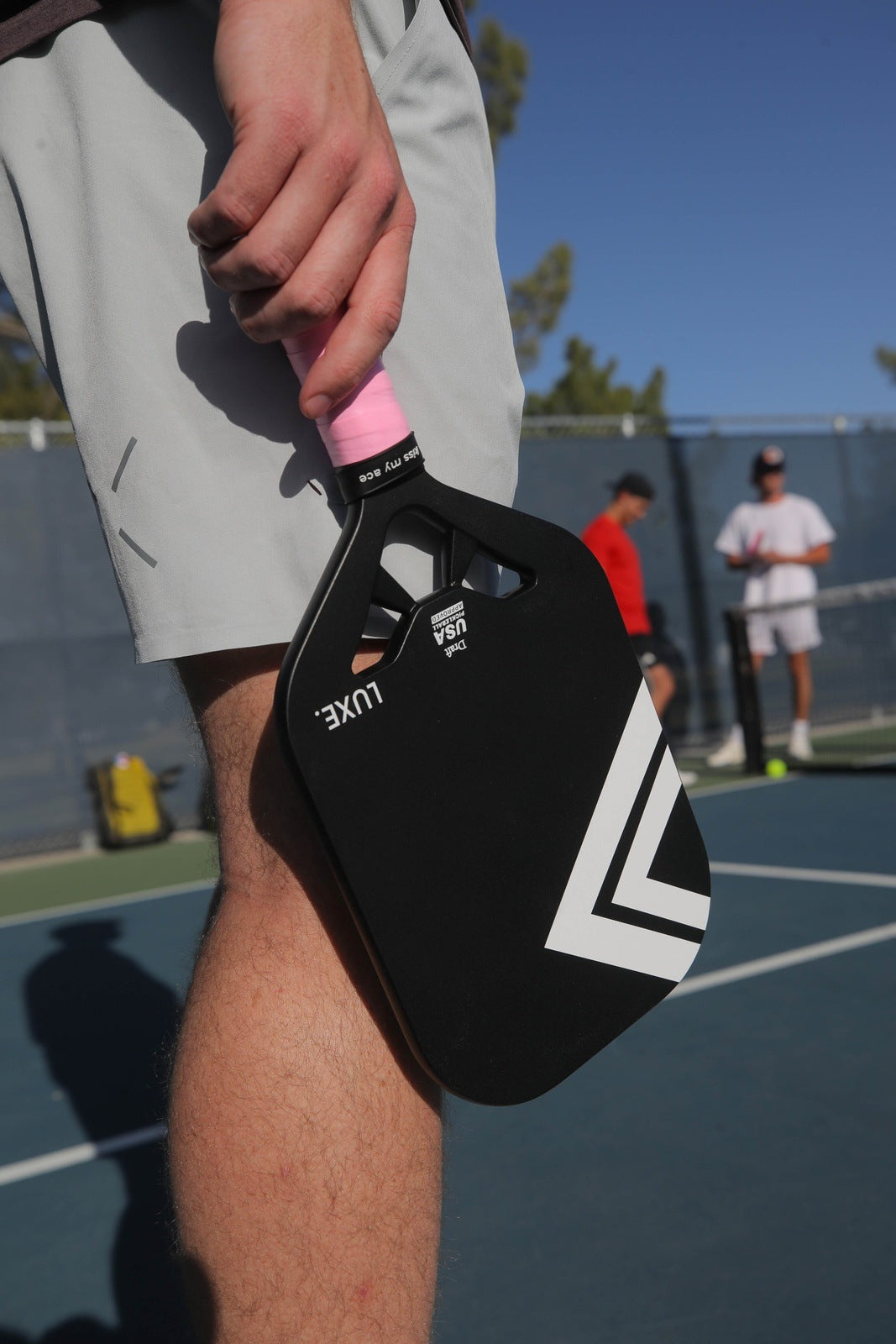 A close-up shows a person holding the LUXE Pickleball Draft paddle, featuring black carbon fiber with white geometric designs and a pink grip, on a blue outdoor court. Two others stand by the net and fence under a clear sky.