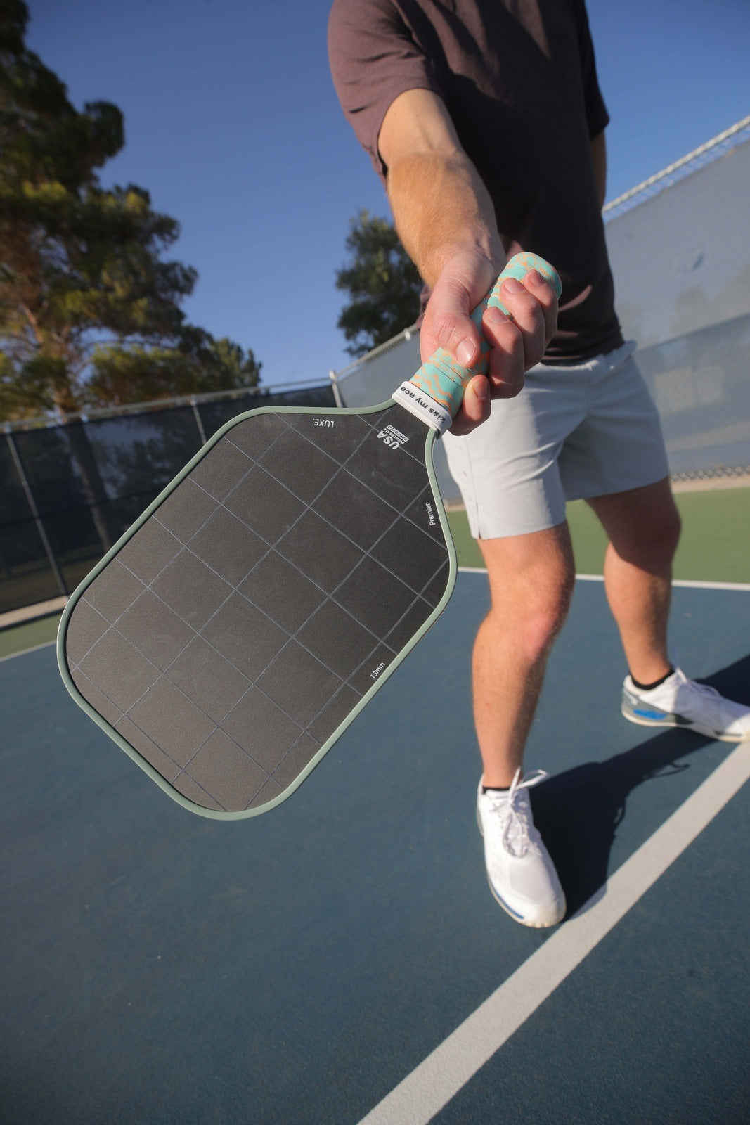 A person in a brown shirt and light shorts stands on an outdoor court, holding a LUXE Pickleball Premier paddle toward the camera. The Premier paddle is in sharp focus, with trees, a fence, and blue sky in the background.