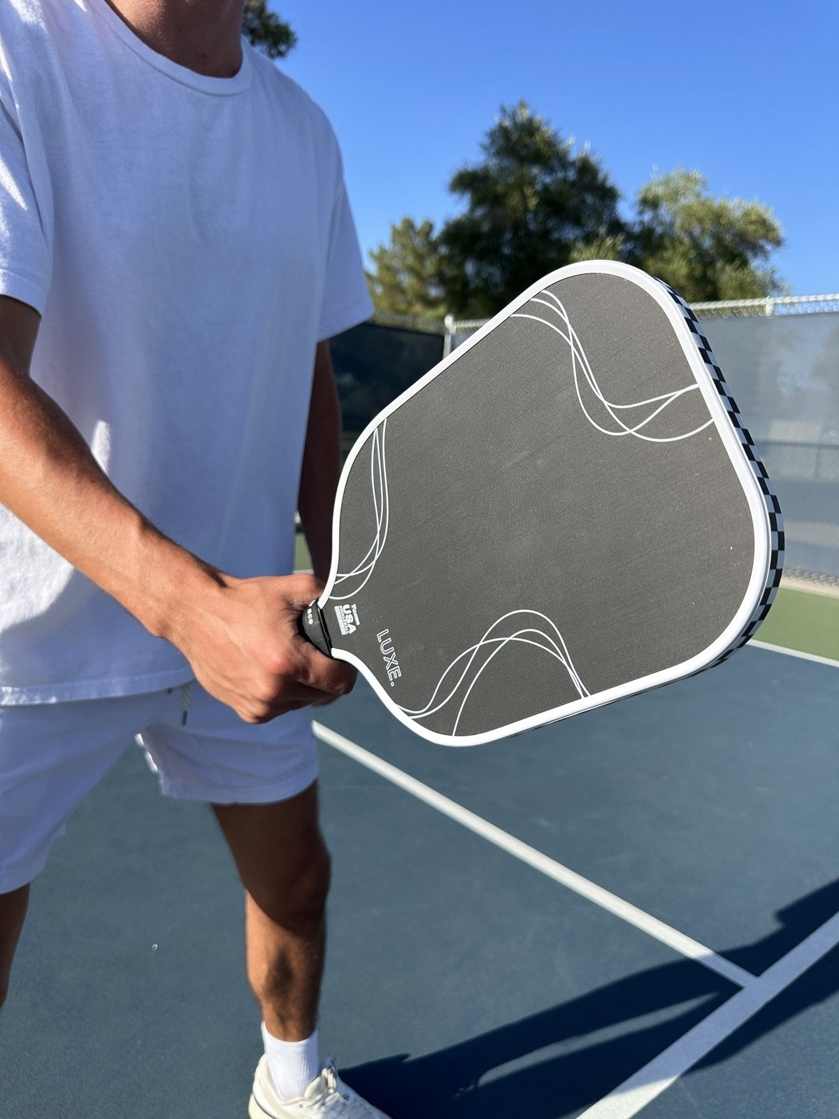 A person in white stands on a pickleball court holding the LUXE Pickleball Finesse carbon fiber paddle close to the camera, with trees and a blue sky in the background. Their face is not visible.