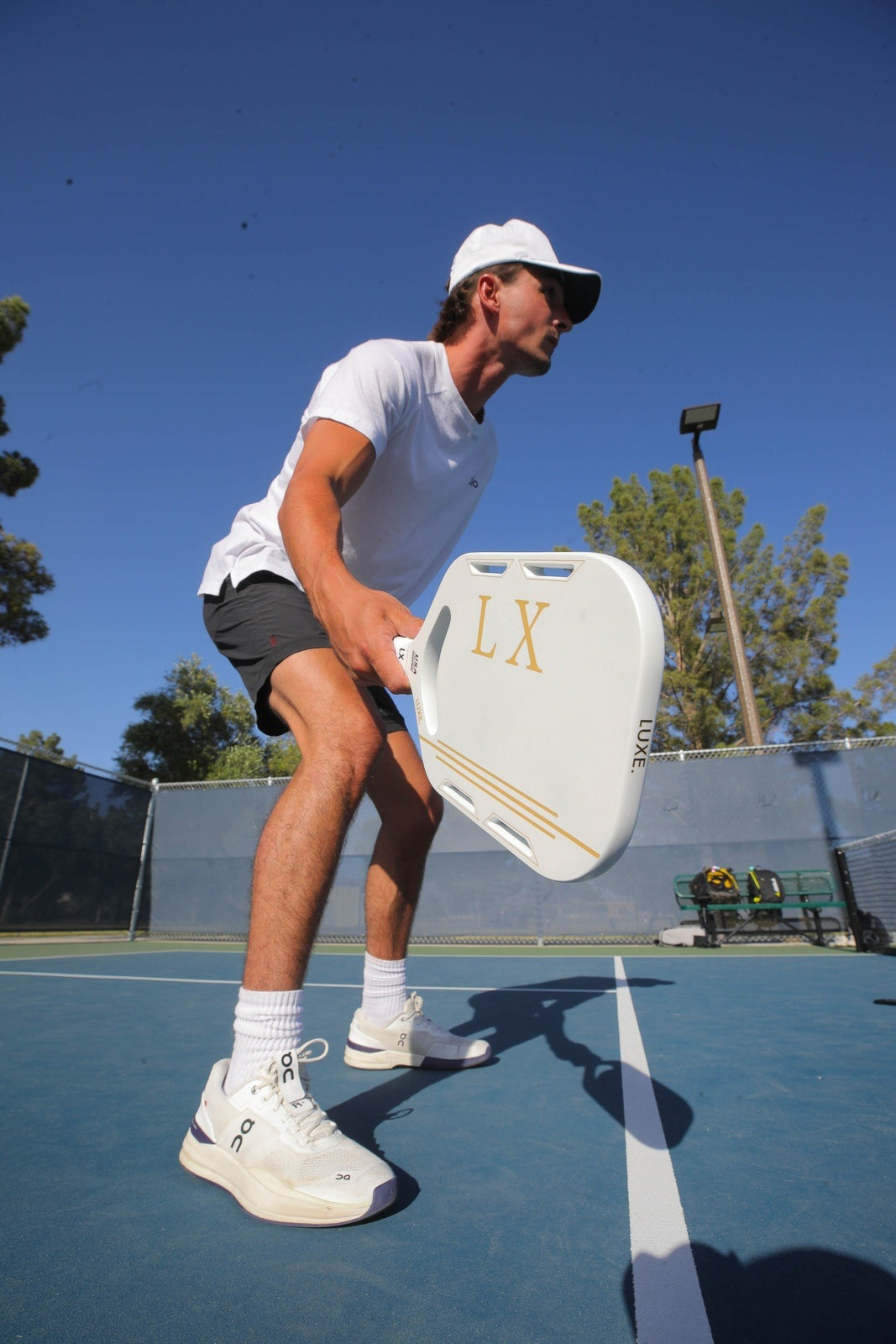 A man in a white cap, sunglasses, and white shirt plays pickleball outdoors with the LX paddle by LUXE Pickleball, bending forward beneath a sunny blue sky and trees.