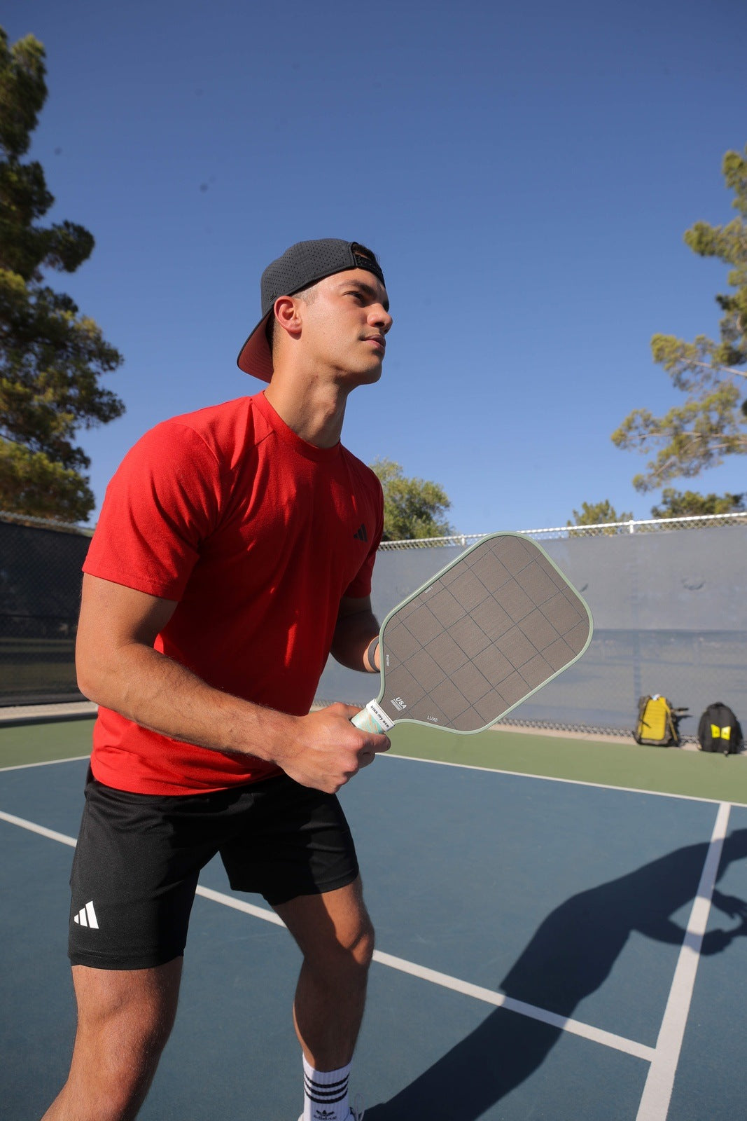 A young man in a red shirt and black shorts stands on an outdoor court, holding the LUXE Pickleball Premier raw carbon fiber paddle. Sunlight highlights two yellow bags by the fence, with trees and a blue sky behind him.