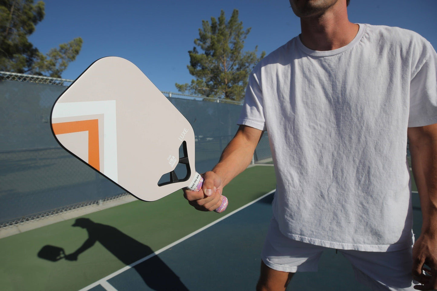 A person in a white T-shirt and shorts holds the LUXE Pickleball Draft paddle—beige with geometric orange and brown lines and an open throat design—on an outdoor court near a chain-link fence, with trees and blue sky in the background.