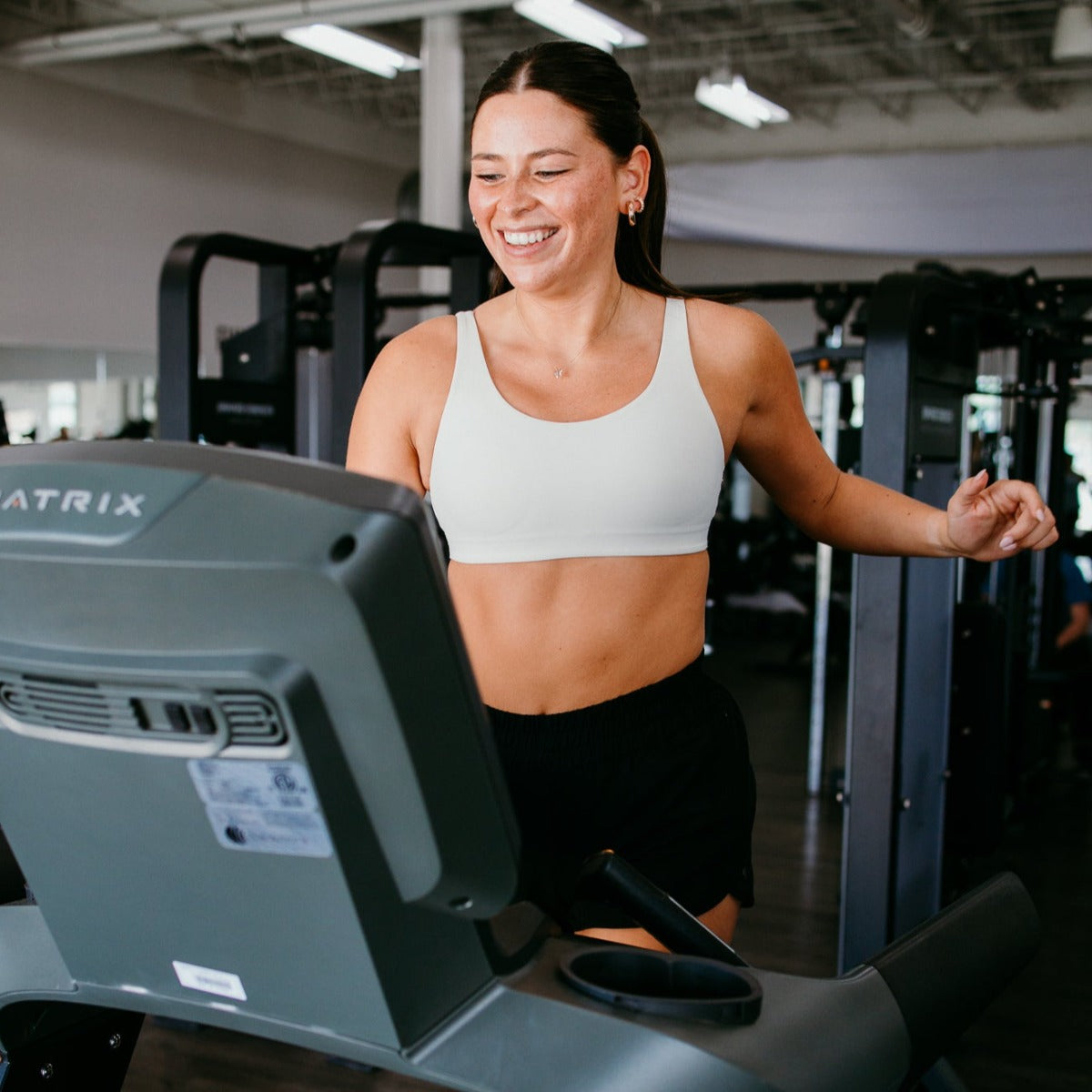 A woman with brown hair tied back smiles as she runs on a treadmill, wearing a white sports bra and DYM Athletics' Sprint High-Rise Lined Short | 3" in black. Small hoop earrings, bright gym equipment, and mirrors fill the background.