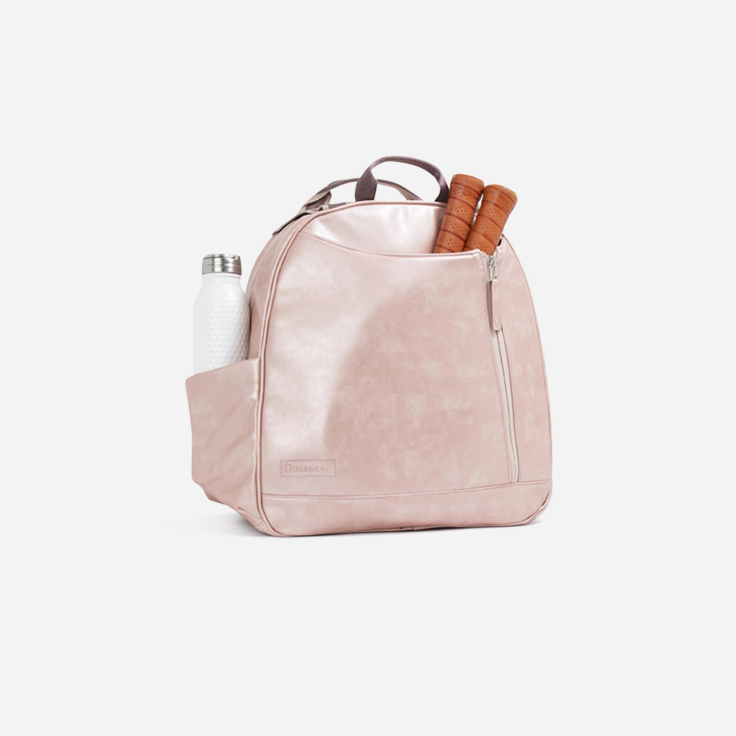 The Doubletake MELBOURNE Pickleball Backpack & Tote is a pale pink, water-resistant bag with a top handle and front zip pocket. Two racket handles peek out from the main compartment; a white bottle sits in the side pocket against a plain white background.