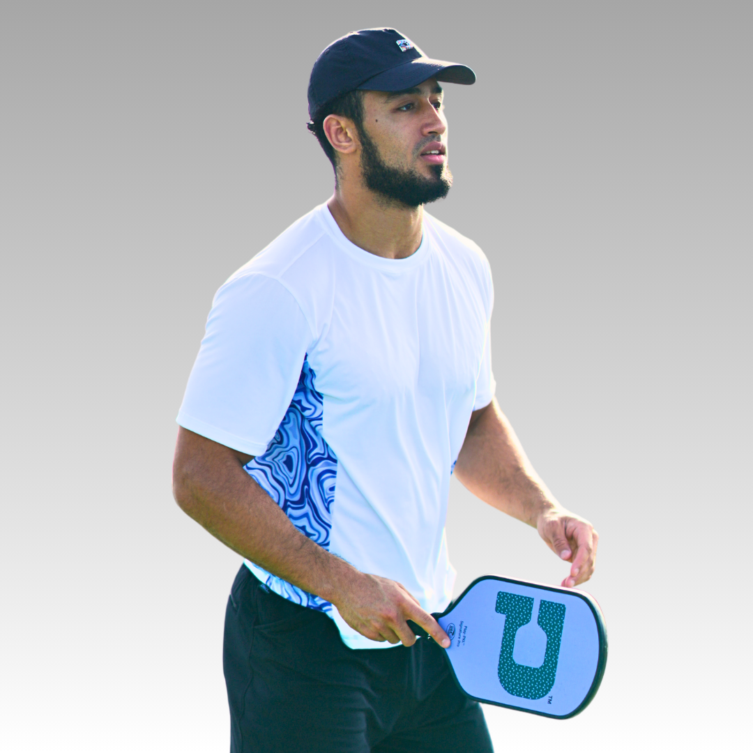 A bearded man in the Playly Store’s Playly - Warrior Tee 2.0, black shorts, and a black cap holds a pickleball paddle, appearing focused against a plain gray gradient background.