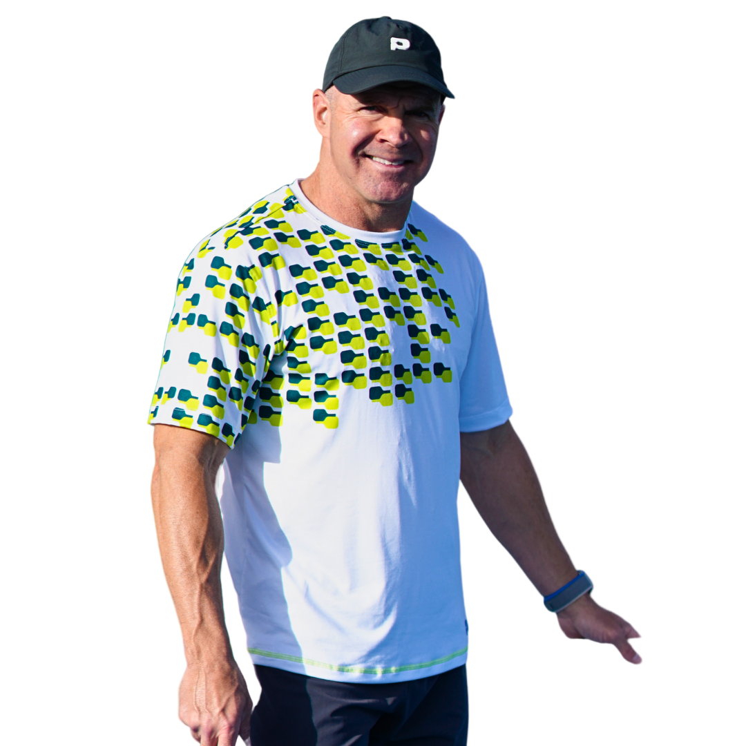 A smiling man in a black cap and Playly - Warrior Tee 2.0 from Playly Store, featuring blue and green geometric patterns and modern moisture-wicking fabric, stands outdoors wearing a fitness tracker. The background is white.