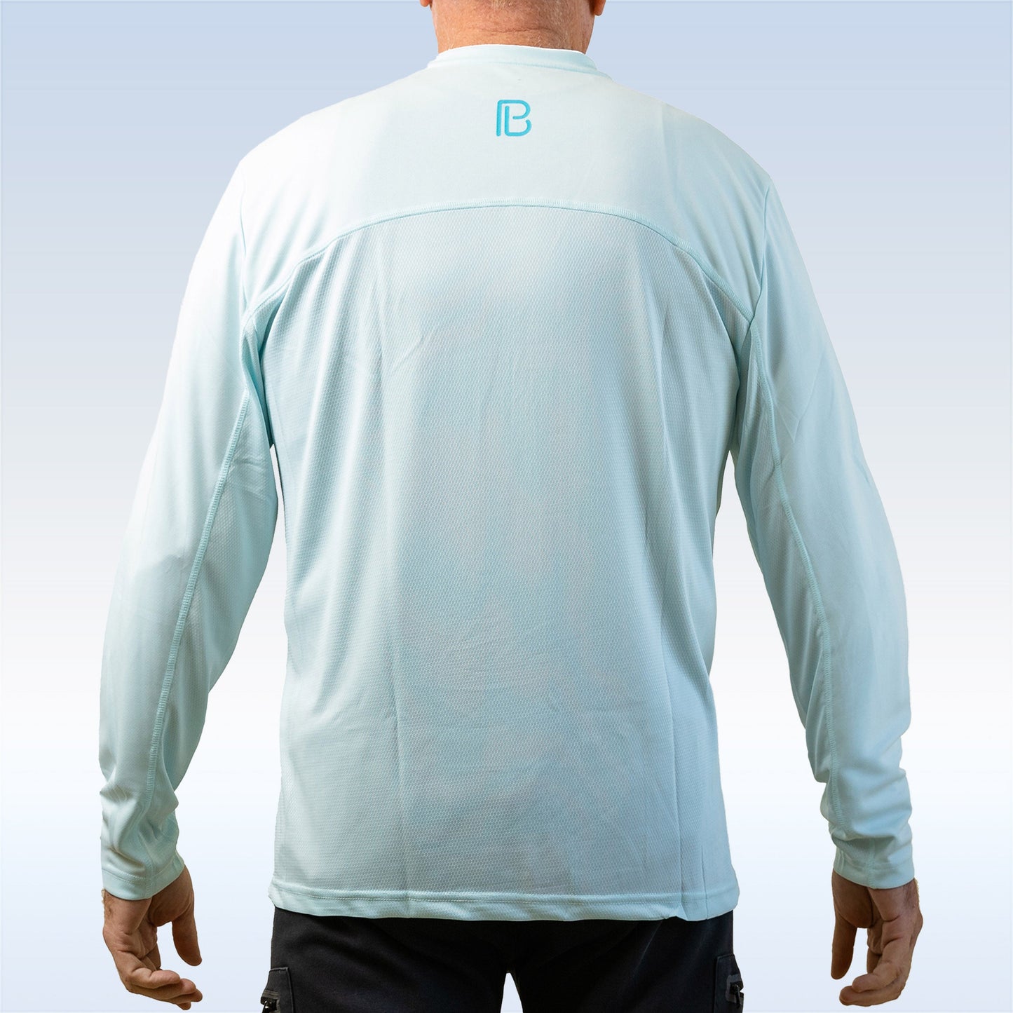 A man wearing the PB Men's Aqua Long Sleeve Tee by Pickleball Bella stands with his back to the camera. The light blue shirt features a blue “B” logo at the upper back and offers SPF 30+ sun protection, set against a pale gradient background.