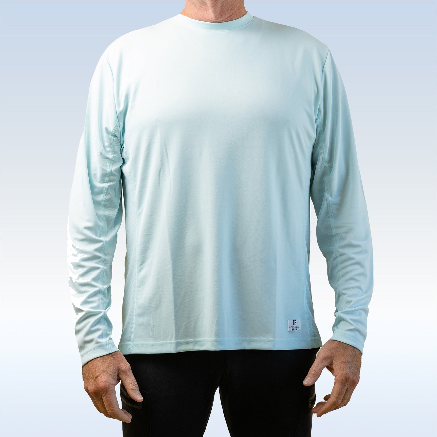 A man wears a PB Men's Aqua Long Sleeve Tee by Pickleball Bella, facing forward against a pale gradient background. The light blue shirt offers SPF 30+ sun protection and moisture wicking. Only his torso and arms are visible; the shirt is pattern-free.
