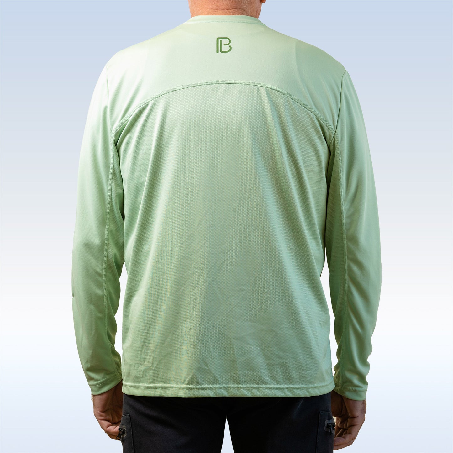 A person wearing a Pickleball Bella PB Men's Smoke Green Long Sleeve Tee, featuring a subtle logo below the collar, stands with their back to a gradient light blue and white background. This SPF 30+ shirt combines style and sun protection.