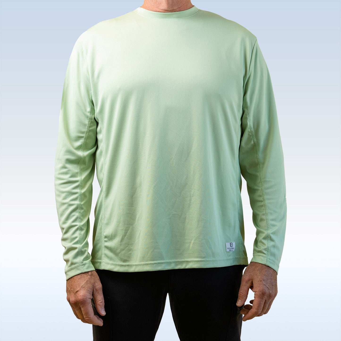 A man wears a PB Men's Smoke Green Long Sleeve Tee by Pickleball Bella, featuring a small logo tag near the hem. Only his torso and arms are visible against a soft gradient background, and he pairs it with black pants.
