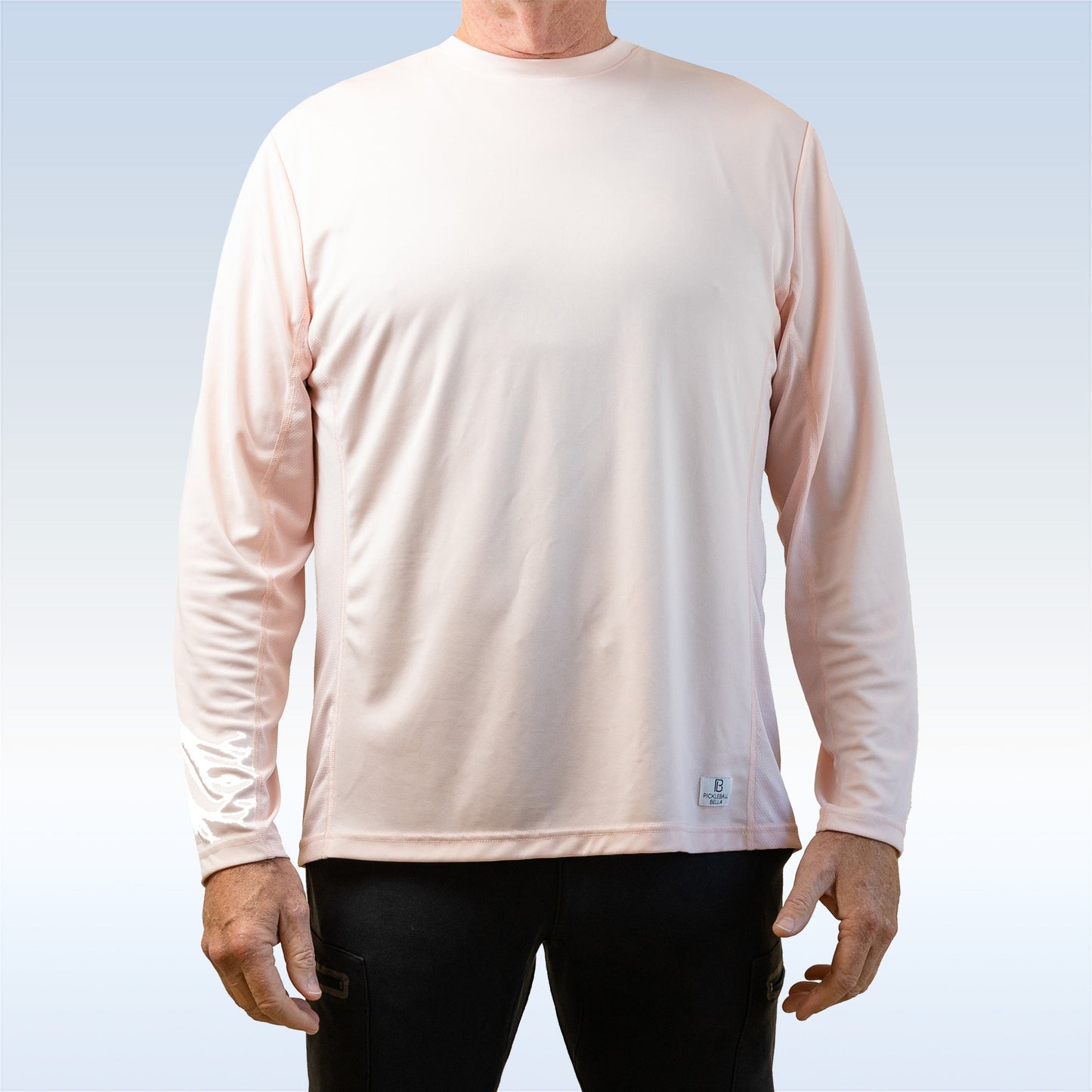 A man faces forward wearing Pickleball Bella's PB Men's Gossamer Pink Long Sleeve Tee and black pants. The plain shirt has a small white tag near the hem, offers moisture wicking, and stands out against a light blue-to-white gradient background.