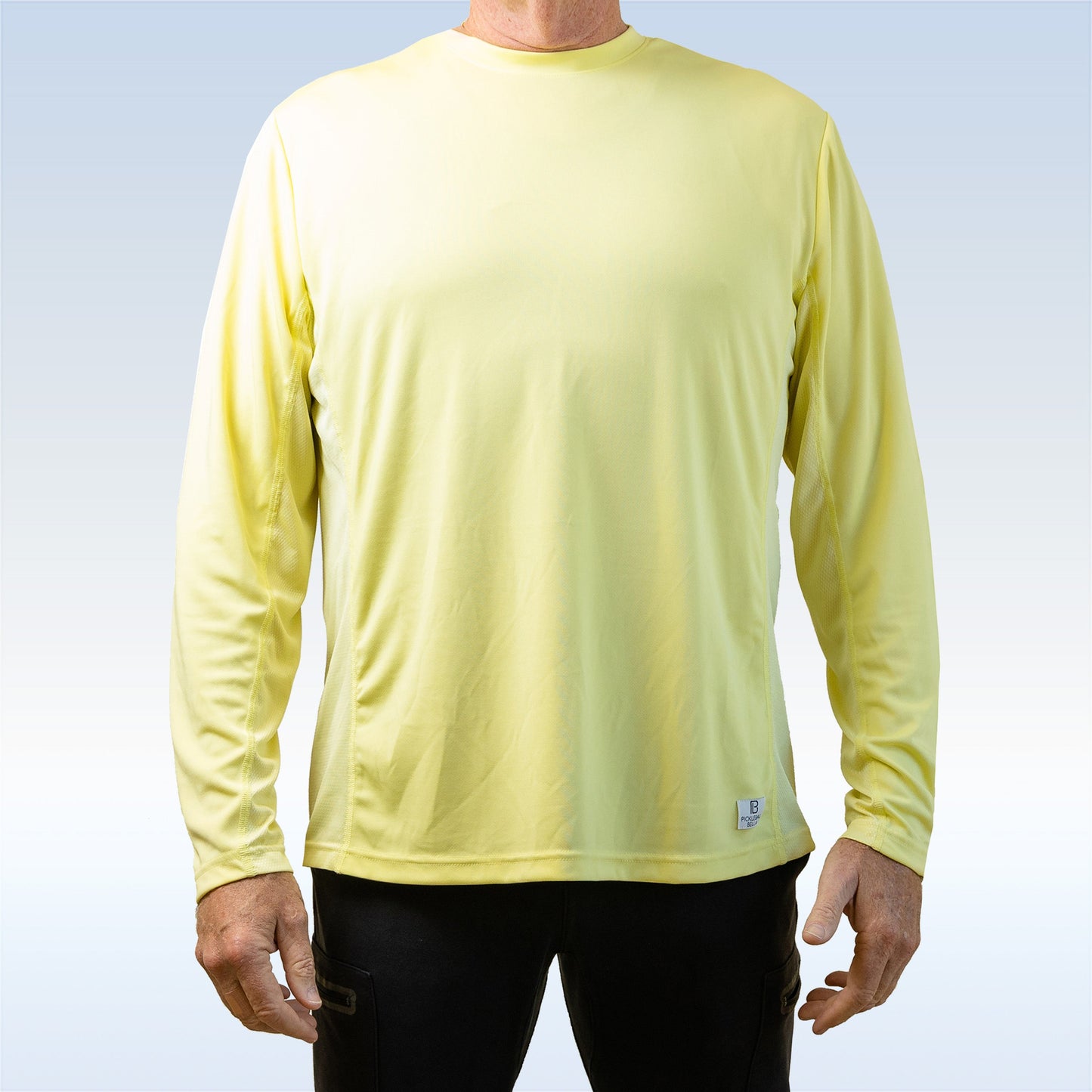 A man wearing a Pickleball Bella PB Men's Elfin Yellow Long Sleeve Tee stands facing forward. The loose, moisture-wicking shirt features SPF 30+ and a small tag near the hem. His hands rest at his sides against a pale blue and white background.