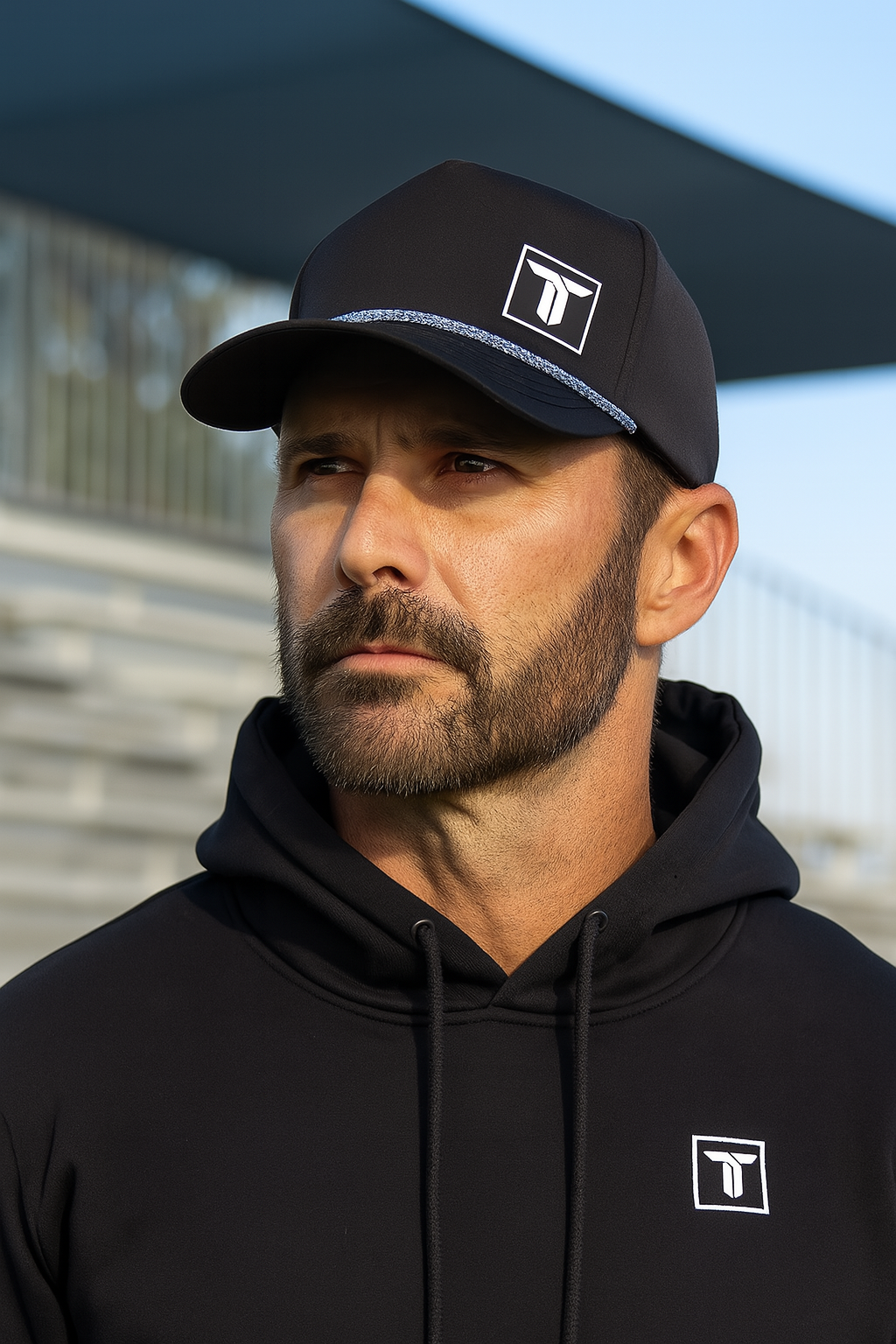 A man with a trimmed beard wears a black Thrive Elite Hoodie by Thrive Pickleball and a matching cap, both with a white geometric logo. He looks serious, glancing to the side near metal bleachers at an outdoor sports venue.