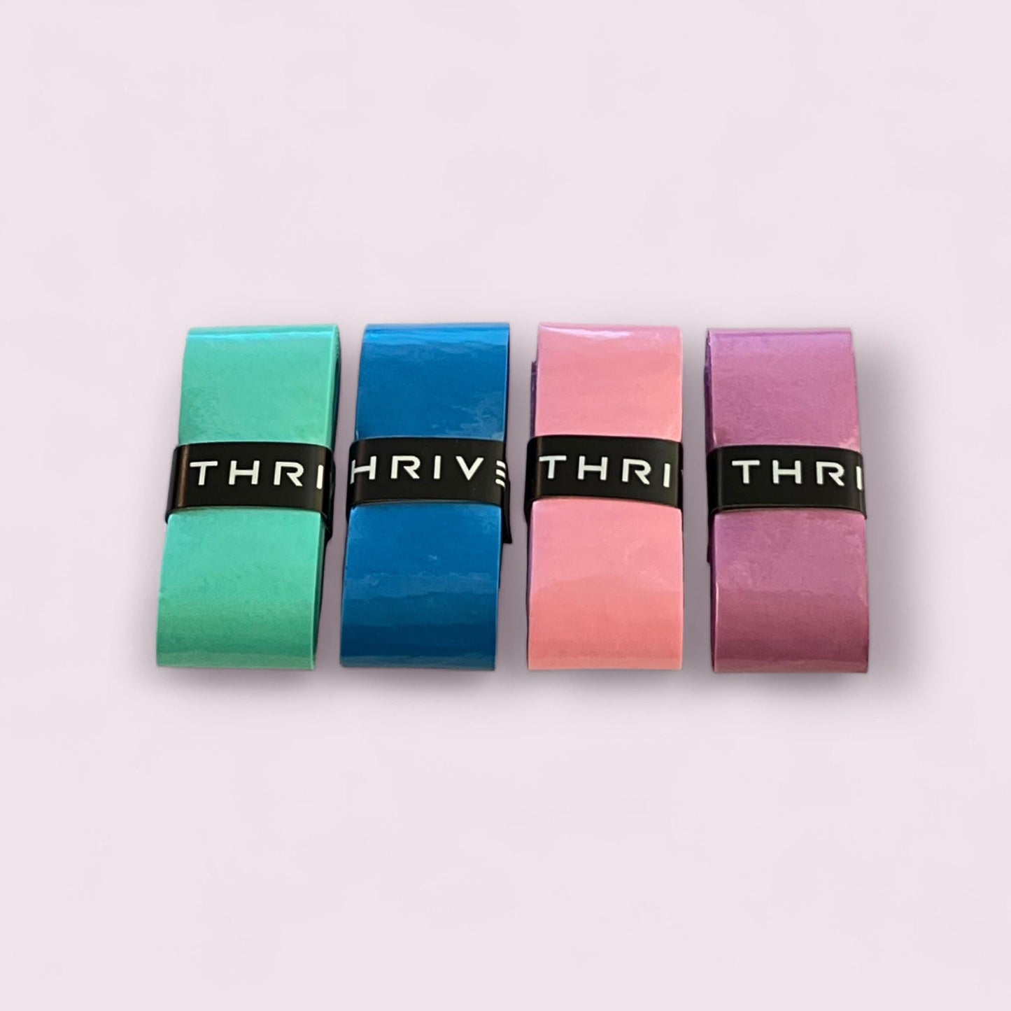 Four rectangular pastel chalks—green, blue, pink, and purple—are lined up on a light pink surface. Each chalk is wrapped with a black “Thrive Pickleball” band, inspired by the super tacky grip of Thrive Pickleball Overgrips.