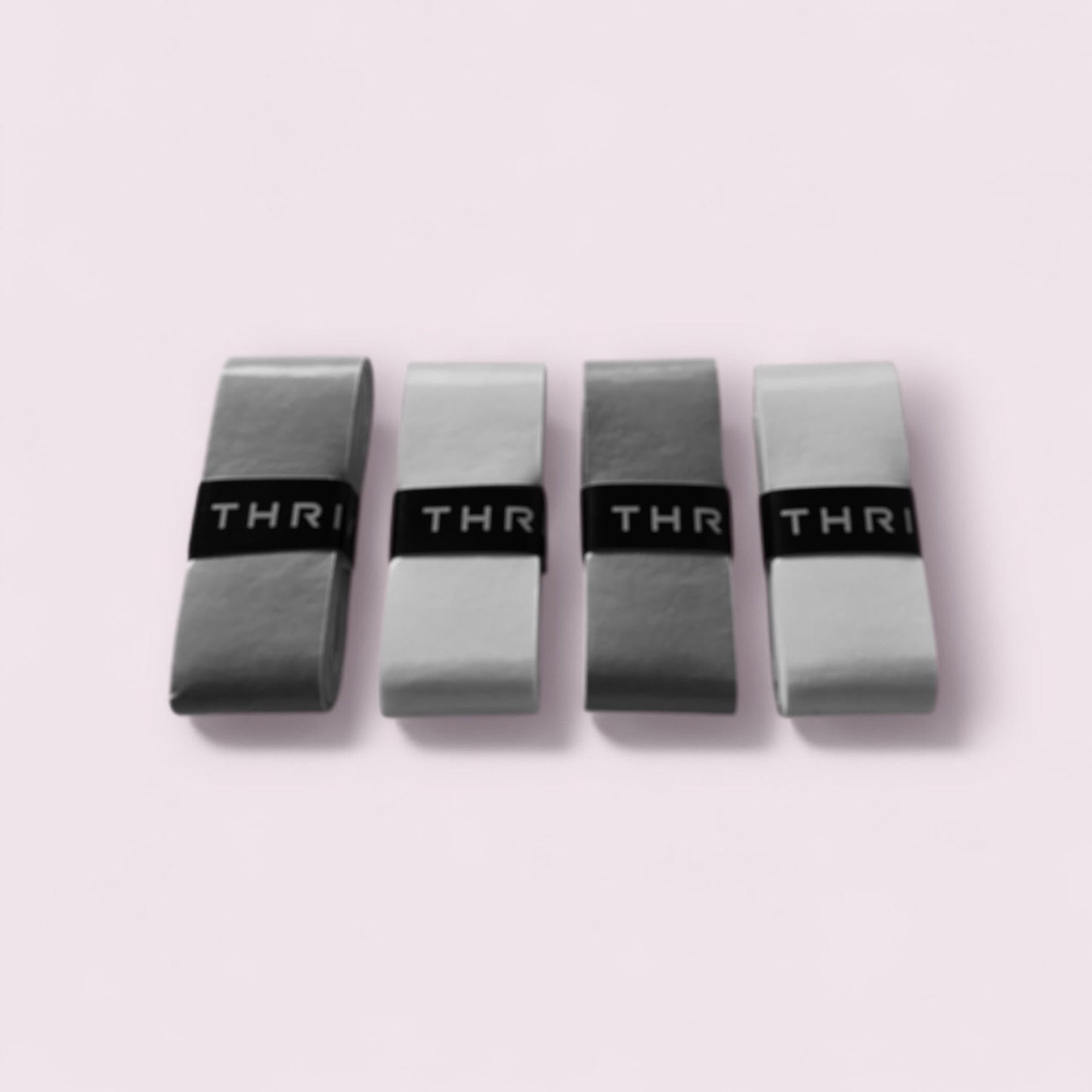Four durable Thrive Pickleball Overgrips (Super Tacky) are neatly rolled and lined up on a light pink background, each secured with a black “THR” band for a sleek, organized look.
