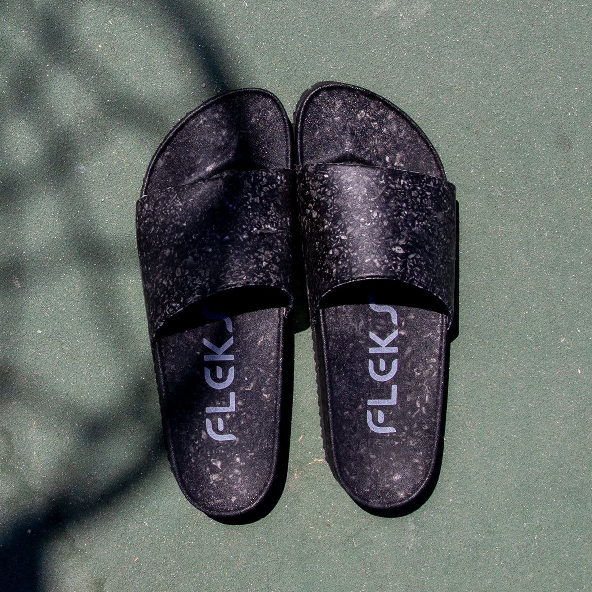 A pair of black, speckled Blumaka Fleks ® East Beach Slides featuring white FLEKS insoles rests side by side on a green surface with diagonal shadows crossing the top left corner, designed to support proper alignment.
