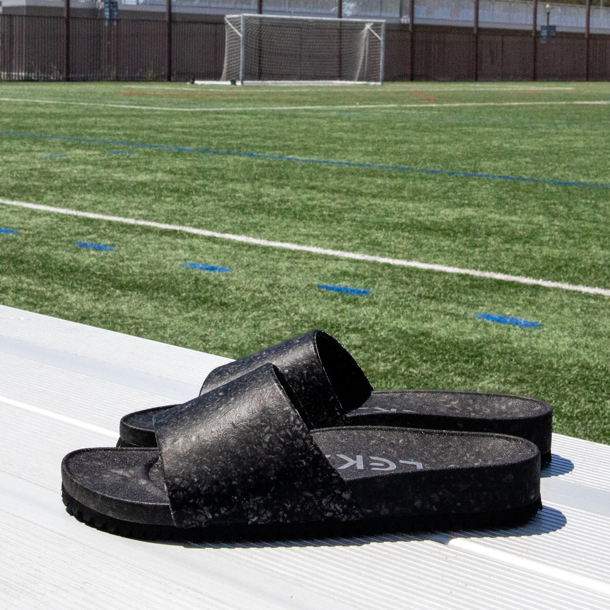 A pair of Blumaka Fleks® East Beach Slide black sandals rest on metal bleachers beside green artificial turf, designed for alignment and recovery, with a goalpost and fence in the background under clear daylight.